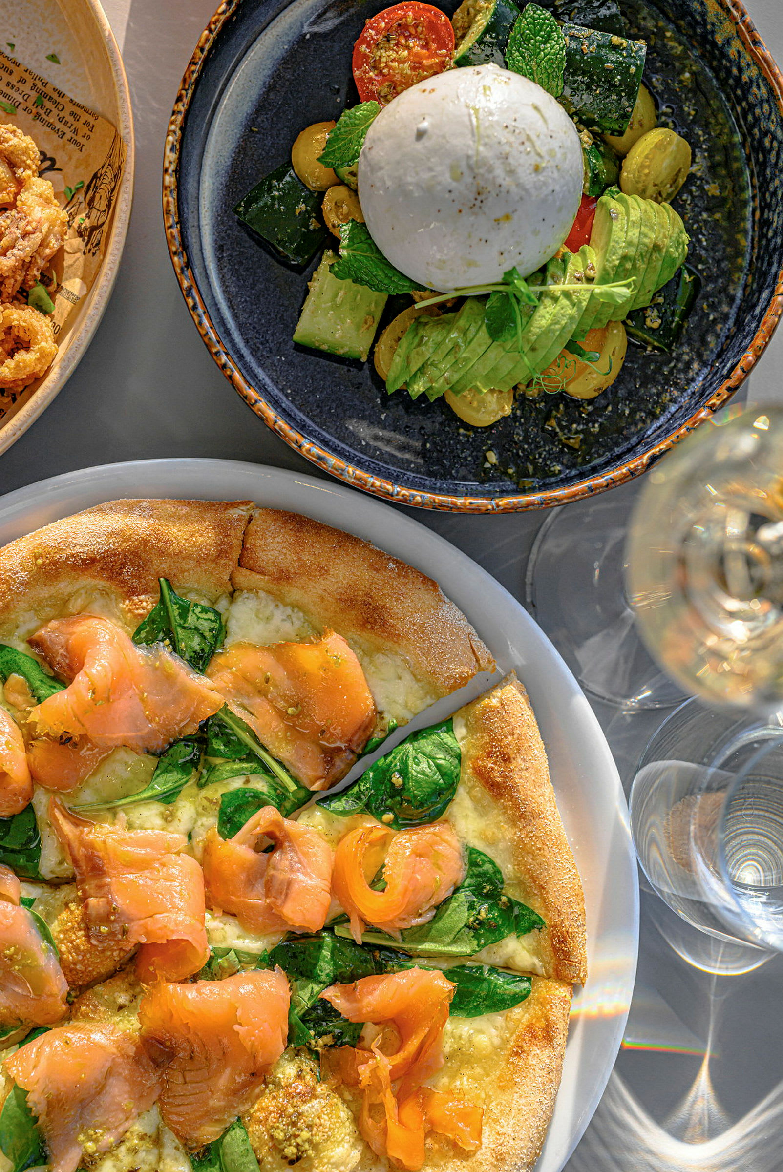En tallerken med burrata, grønnsaker og avokado, samt en pizza med laks og spinat. (AI-generert tekst)