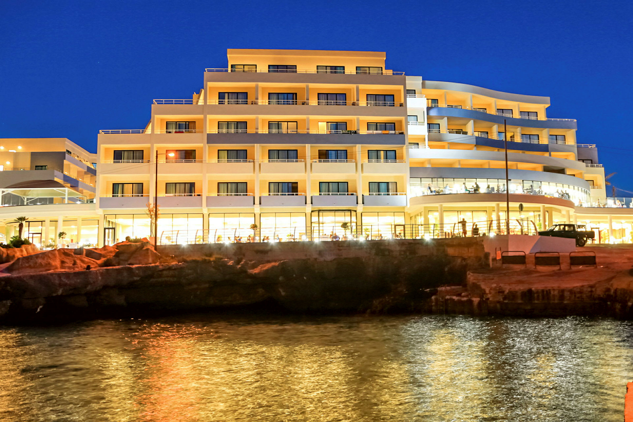 En hotellbygning ved en kyst om kvelden, med lys som reflekteres i vannet. (AI-generert tekst)