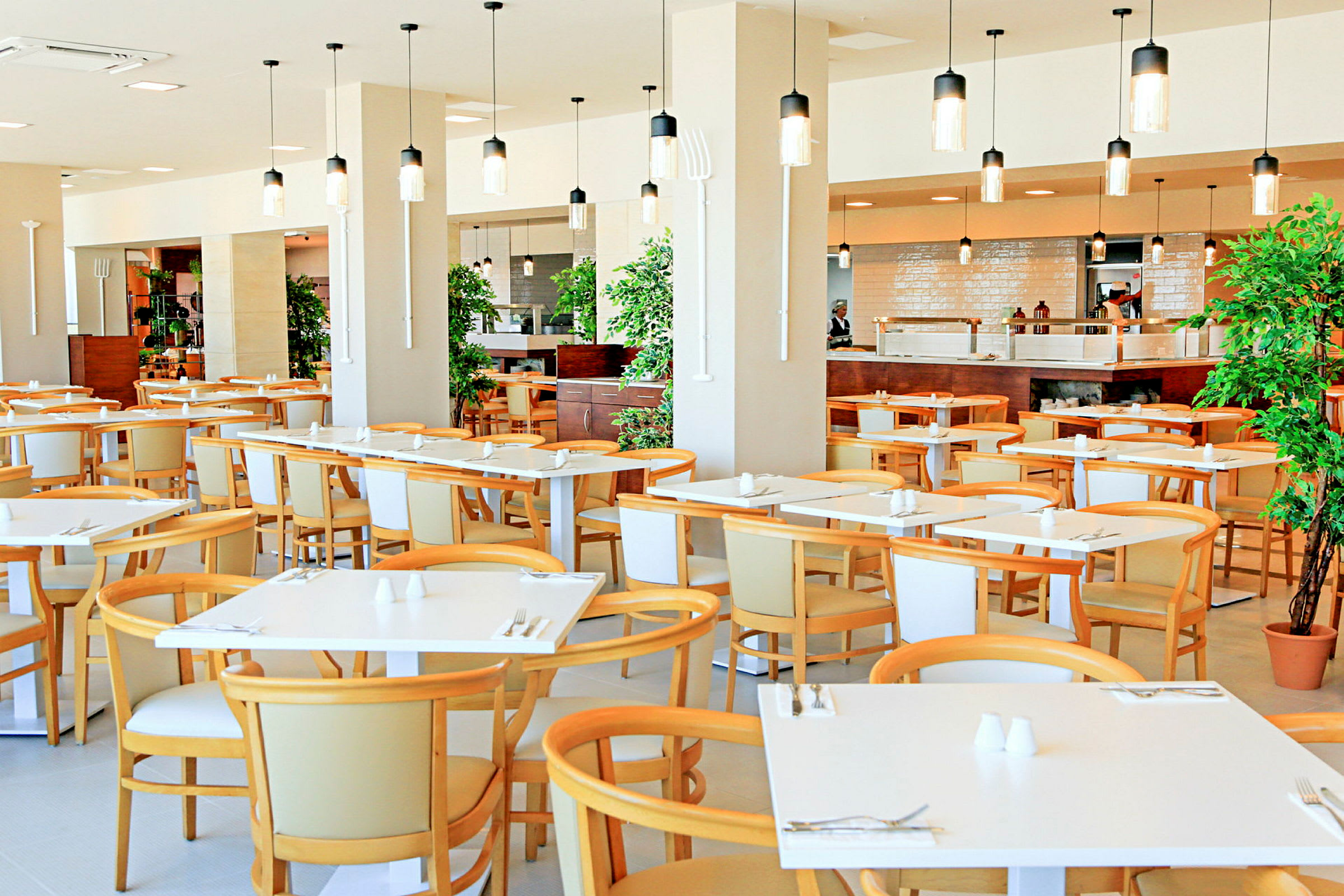 En restaurant med moderne hvite bord og lyse trestoler, dekorert med hengende lamper og planter. (AI-generert tekst)