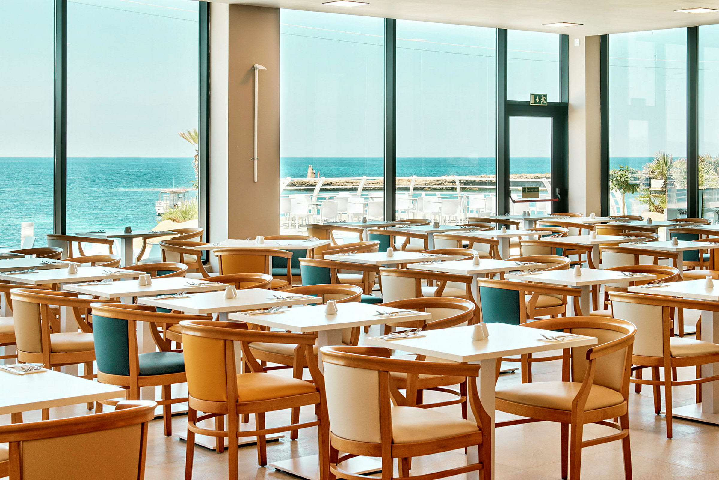 Restaurant med flere bord og stoler, stor vindusutsikt mot havet. (AI-generert tekst)