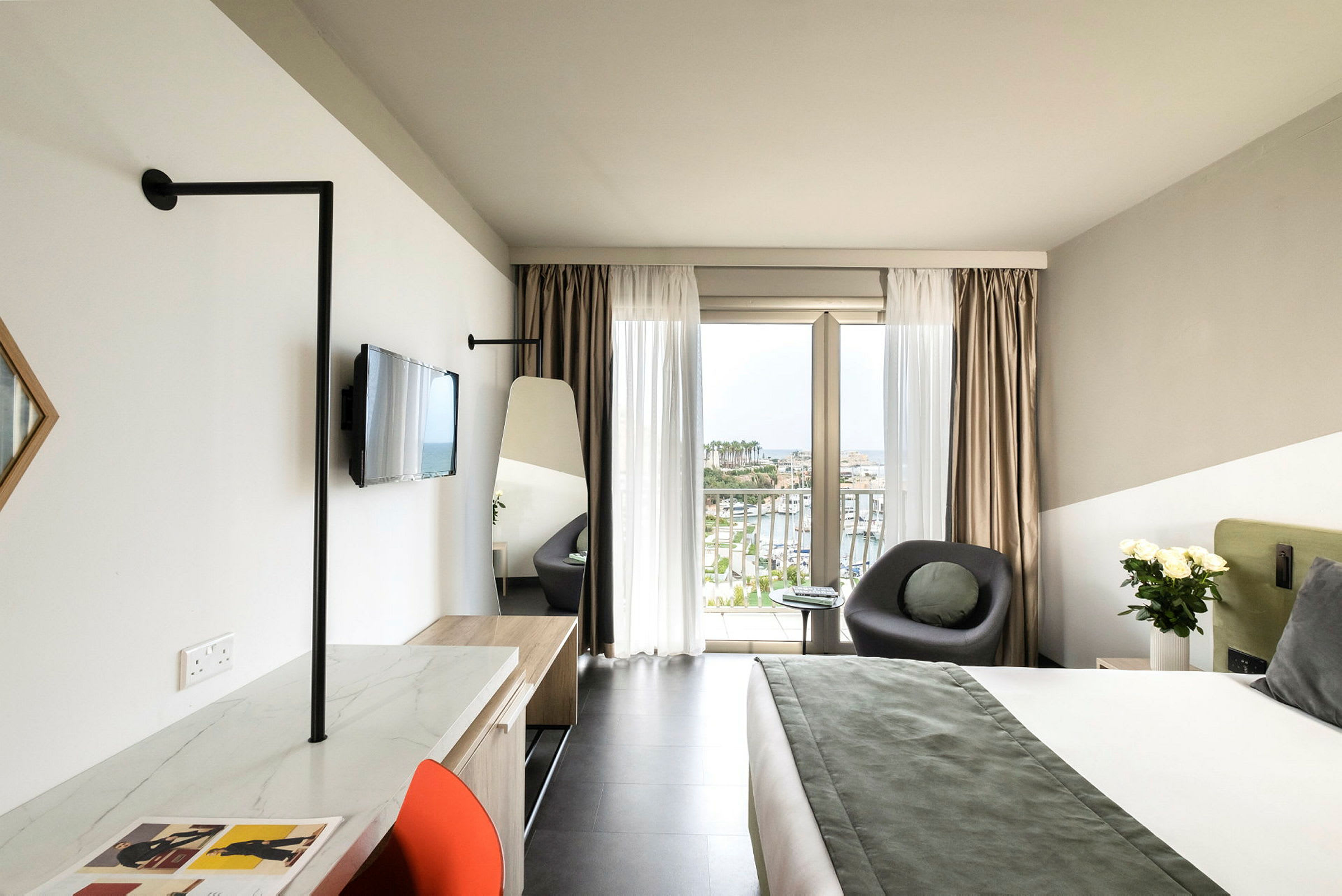 Et moderne hotellrom med en stor seng, en sittegruppe ved et vindu med utsikt over en havn, samt en arbeidsplass med en stol. (AI-generert tekst)