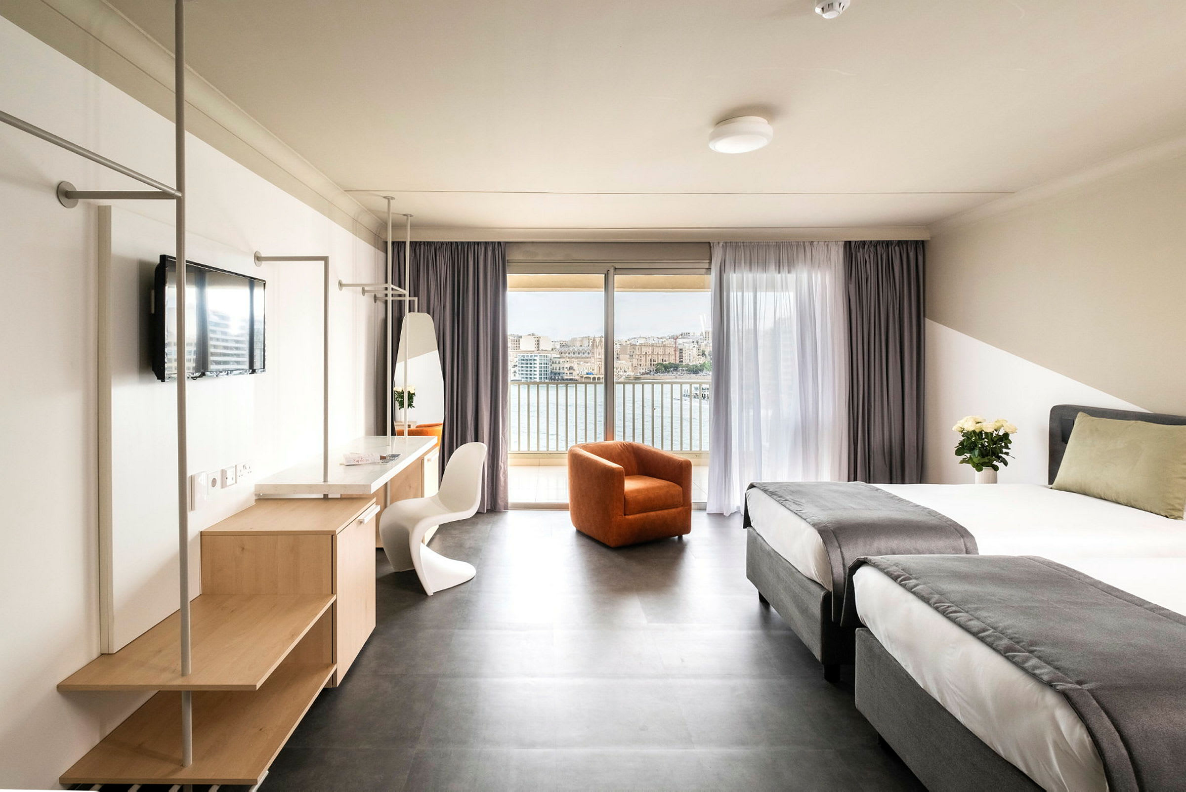 Et hotellrom med to enkeltsenger med grå sengetepper, en oransje lenestol, et skrivebord med speil og TV samt en balkong med utsikt over vann og en by. (AI-generert tekst)