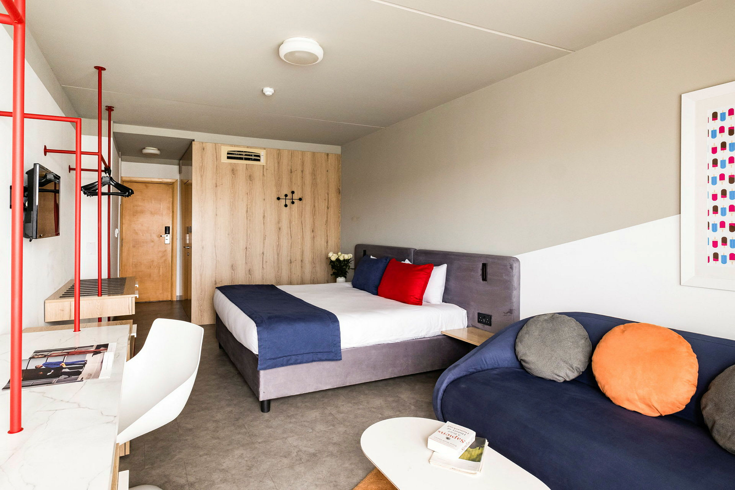 Et hotellrom med en seng med blått sengeteppe og røde og hvite puter, en blå sofa med fargerike puter, samt et trebord og garderobe. (AI-generert tekst)