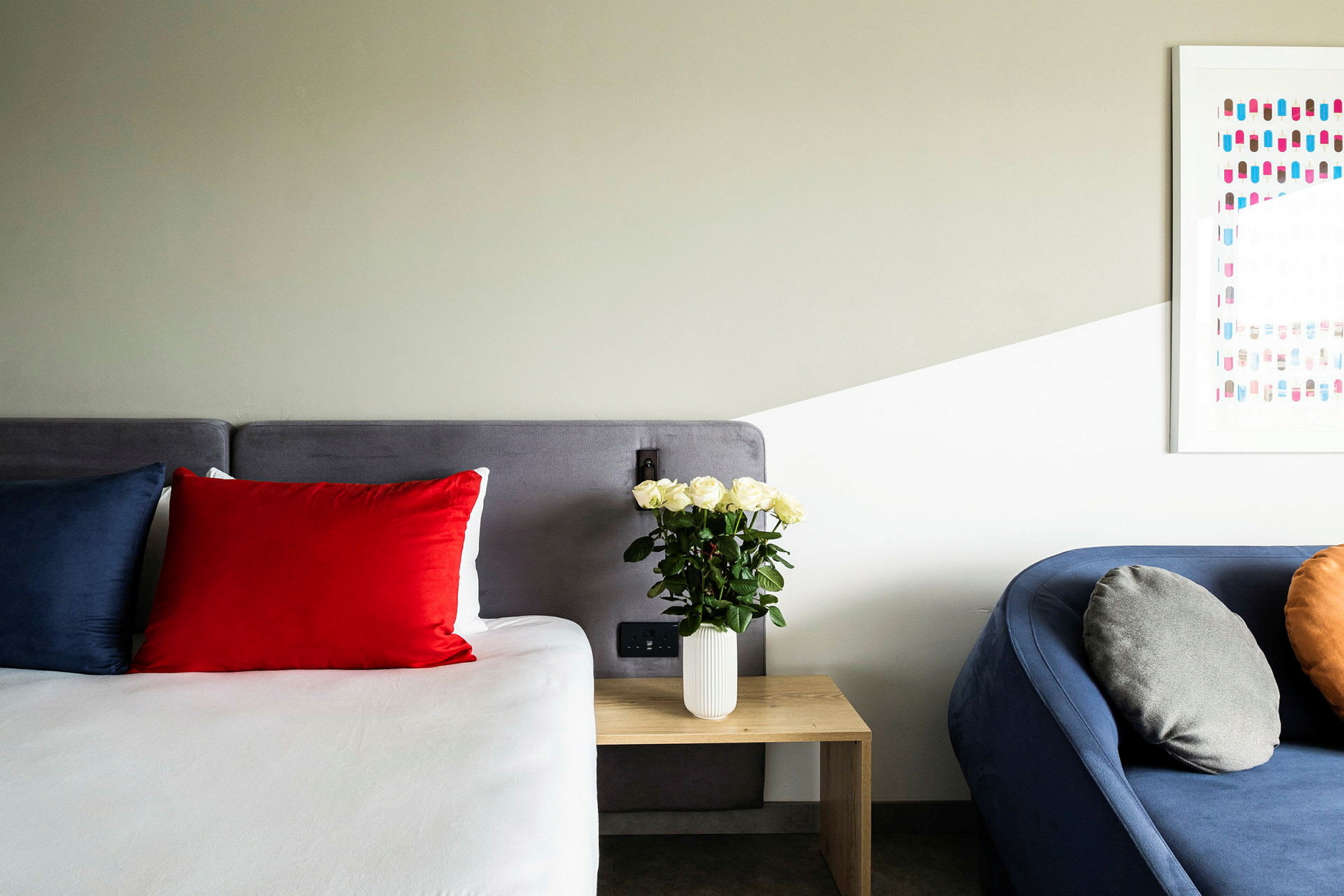 Et rom med en seng med røde og blå puter, et trebord med en vase med hvite roser og en blå sofa med grå og oransje puter. (AI-generert tekst)