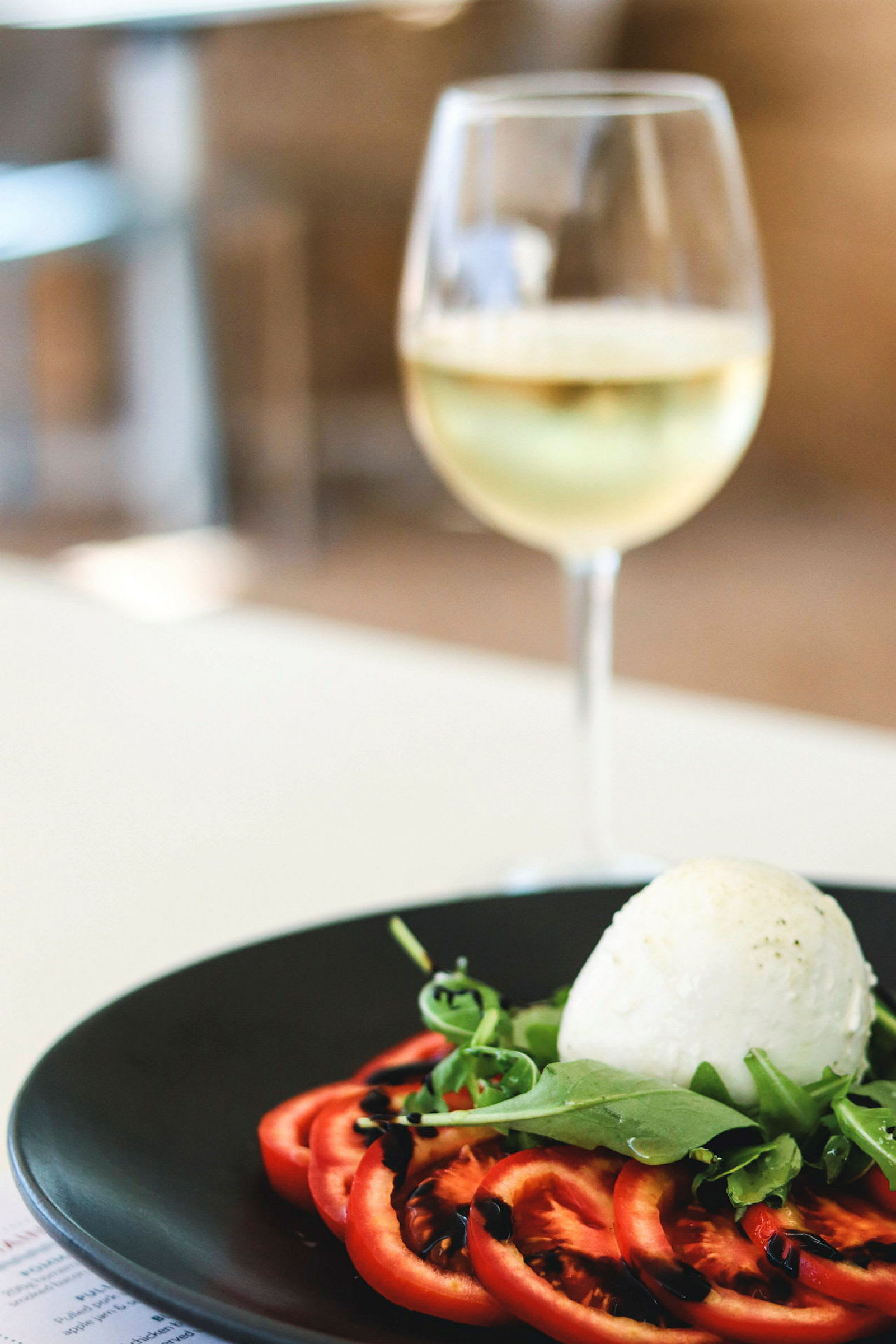 En tallerken med skiver av tomater, ruccola og en ball av mozzarella, toppet med balsamico-krem. I bakgrunnen et glass hvitvin. (AI-generert tekst)