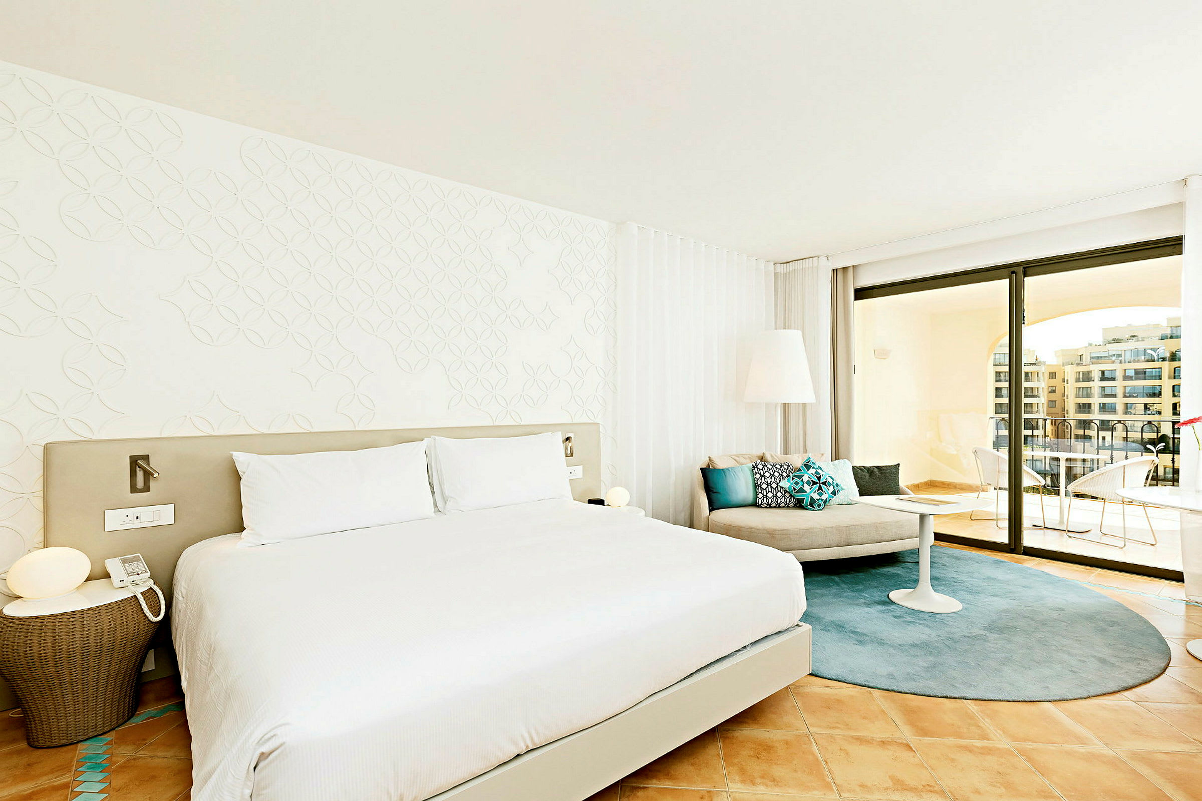 Et moderne hotellrom med en stor seng, lyse vegger og elegant innredning. Rommet har et sitteområde med puter samt en romslig balkong med utsikt. (AI-generert tekst)