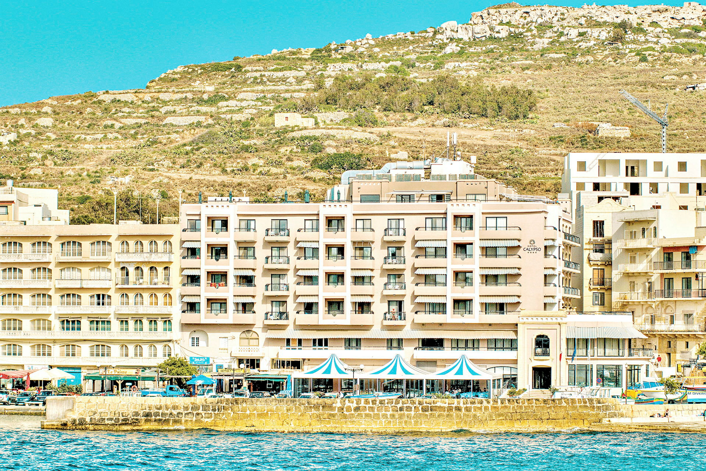 Hotellfasade ved strandpromenaden med kupert terreng i bakgrunnen. (AI-generert tekst)