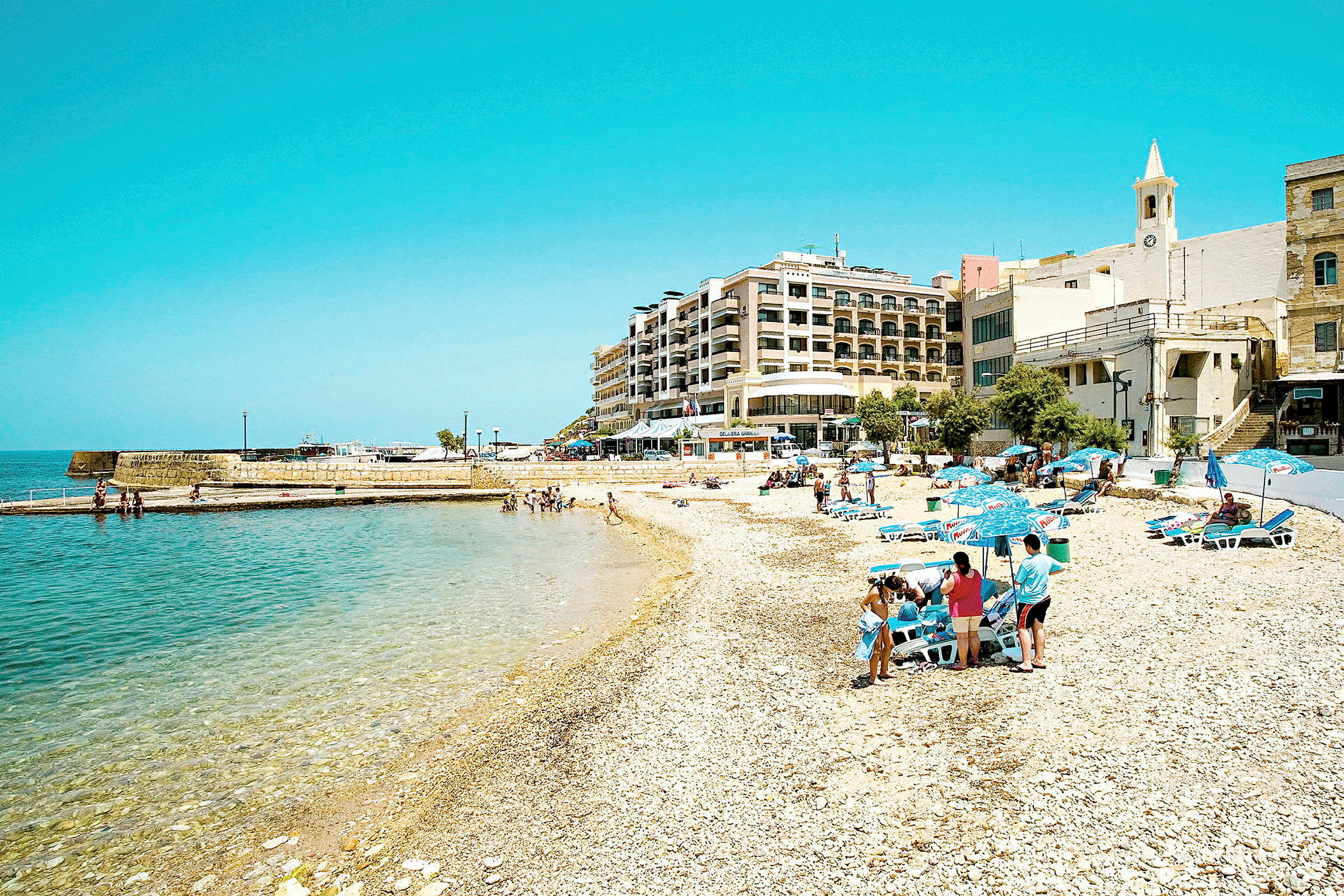 En strand med parasoller og solsenger, en bygning i bakgrunnen. (AI-generert tekst)