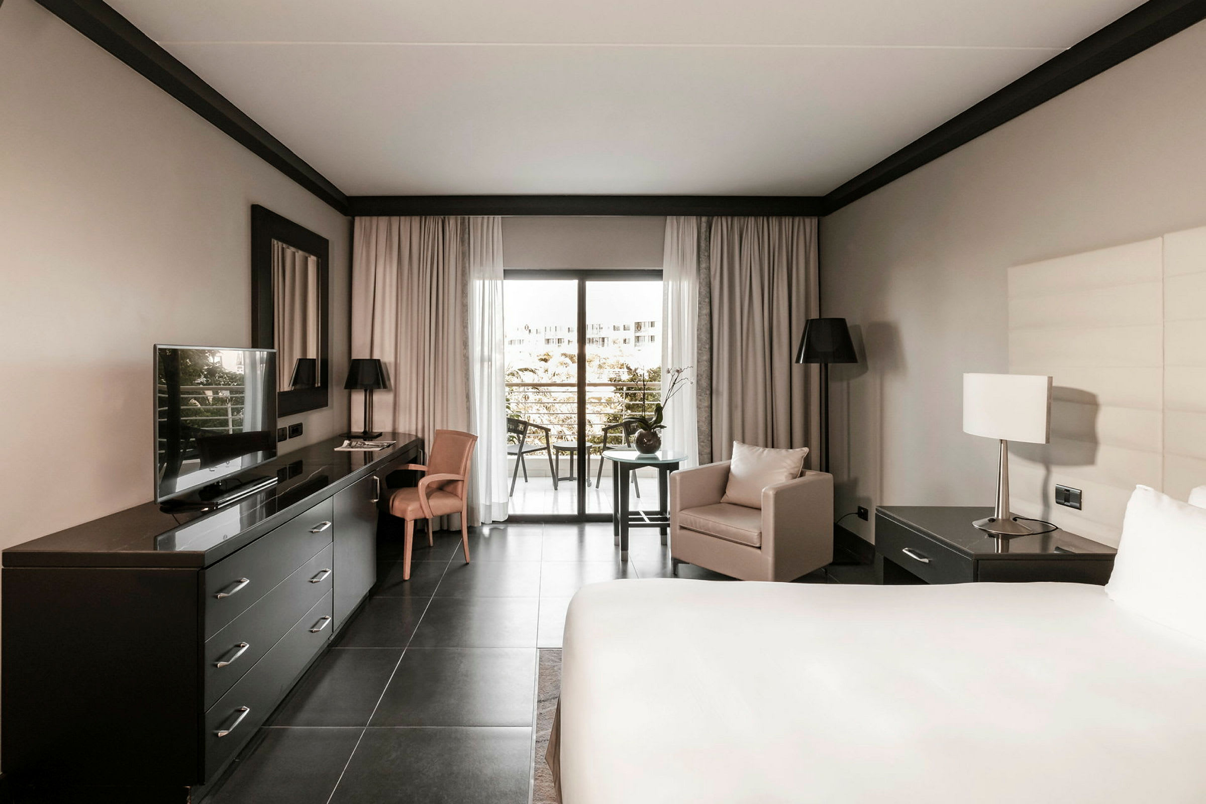Hotellrom med moderne innredning, en stor seng, skrivebord med TV, lenestol og vindu mot en balkong. (AI-generert tekst)