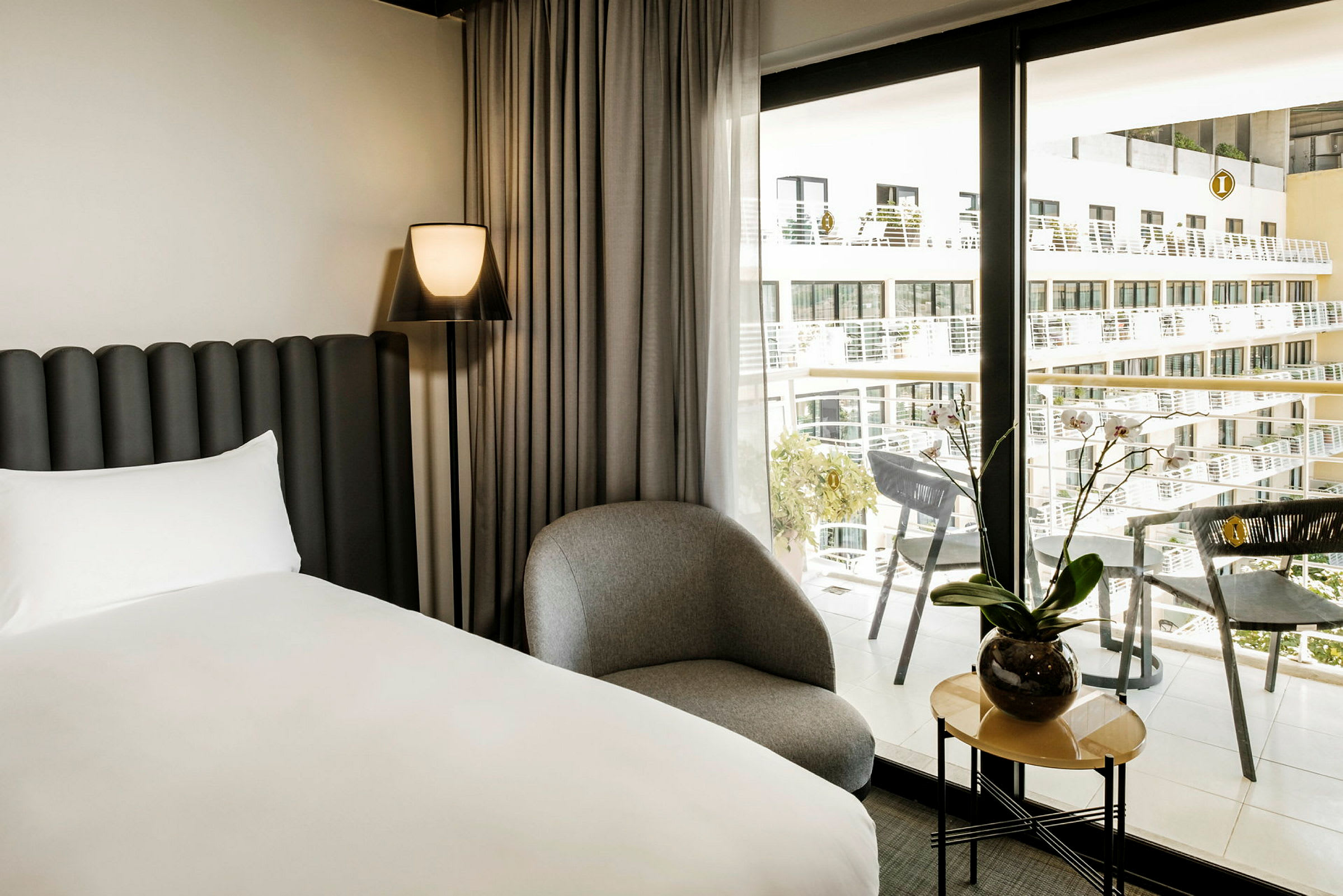Et moderne hotellrom med en seng, en stol og et lite bord med en plante ved siden av et stort vindu med utsikt over balkonger. (AI-generert tekst)