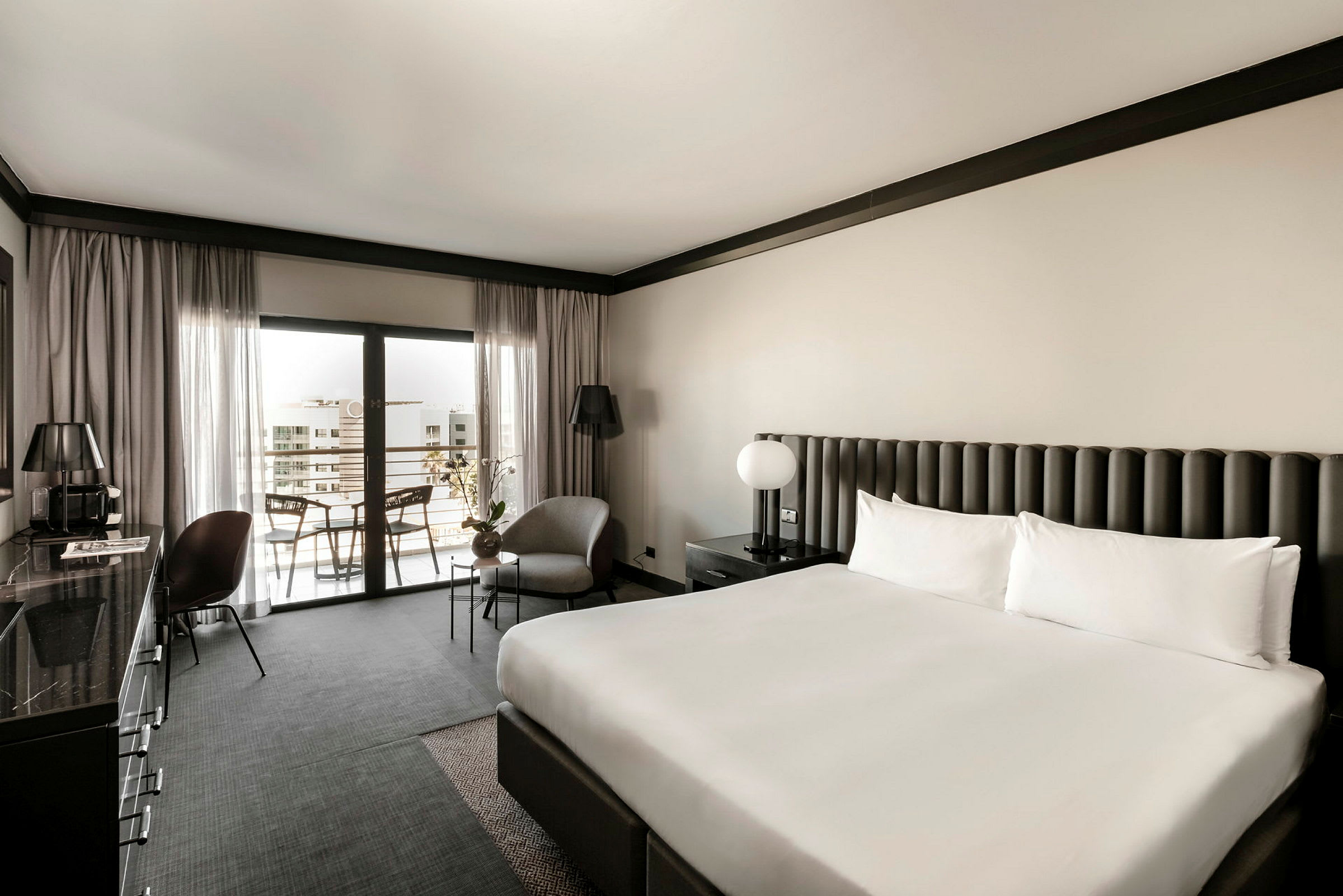 Hotellrom med en stor dobbeltseng, lenestol og bord ved siden av, samt balkong sittplass. Elegant nøytraltonet møbler og innredning. (AI-generert tekst)