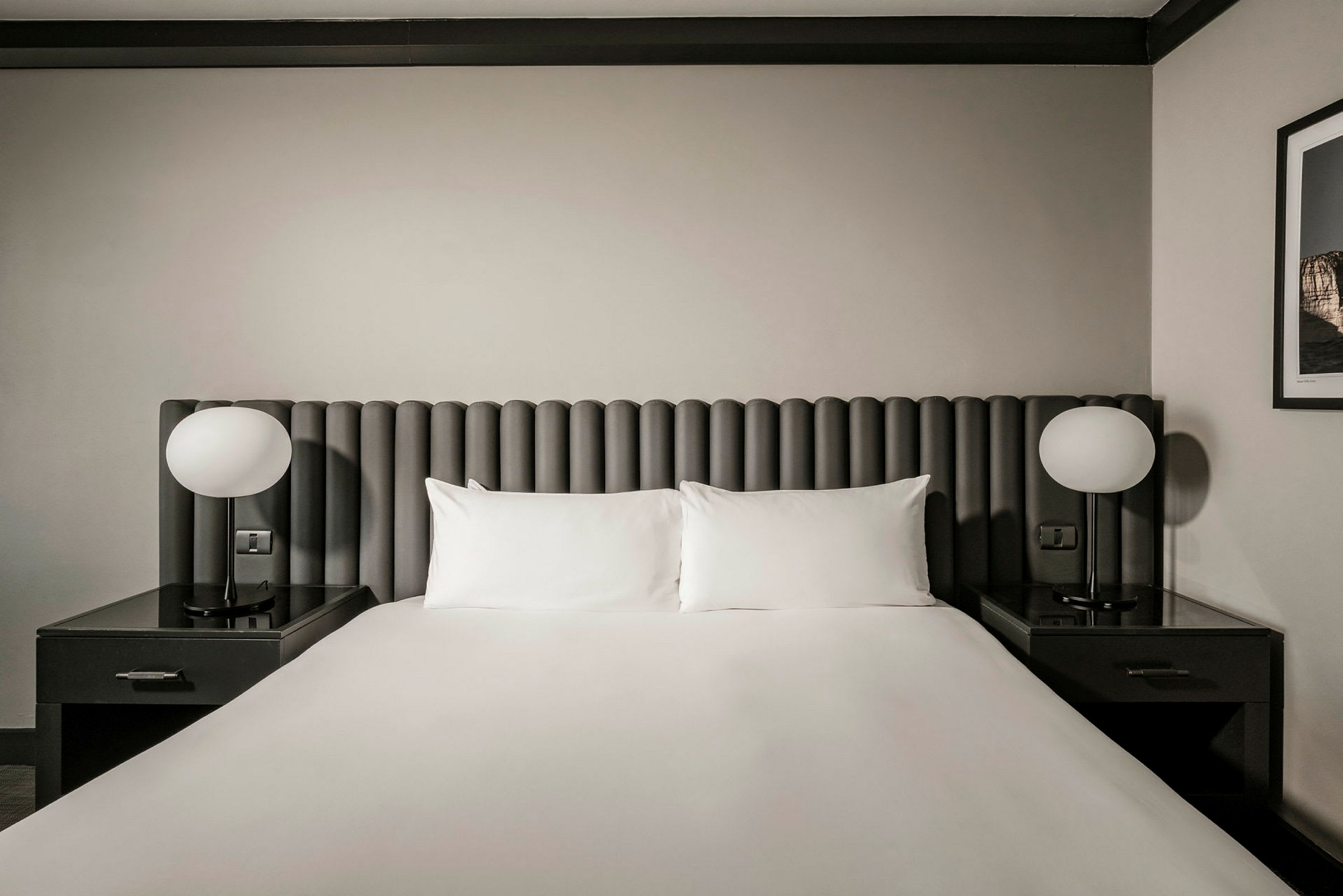 Et hotellrom med en dobbeltseng. Sengen har en grå sengegavl, hvite puter og laken. På hver side er det nattbord med runde lamper. (AI-generert tekst)