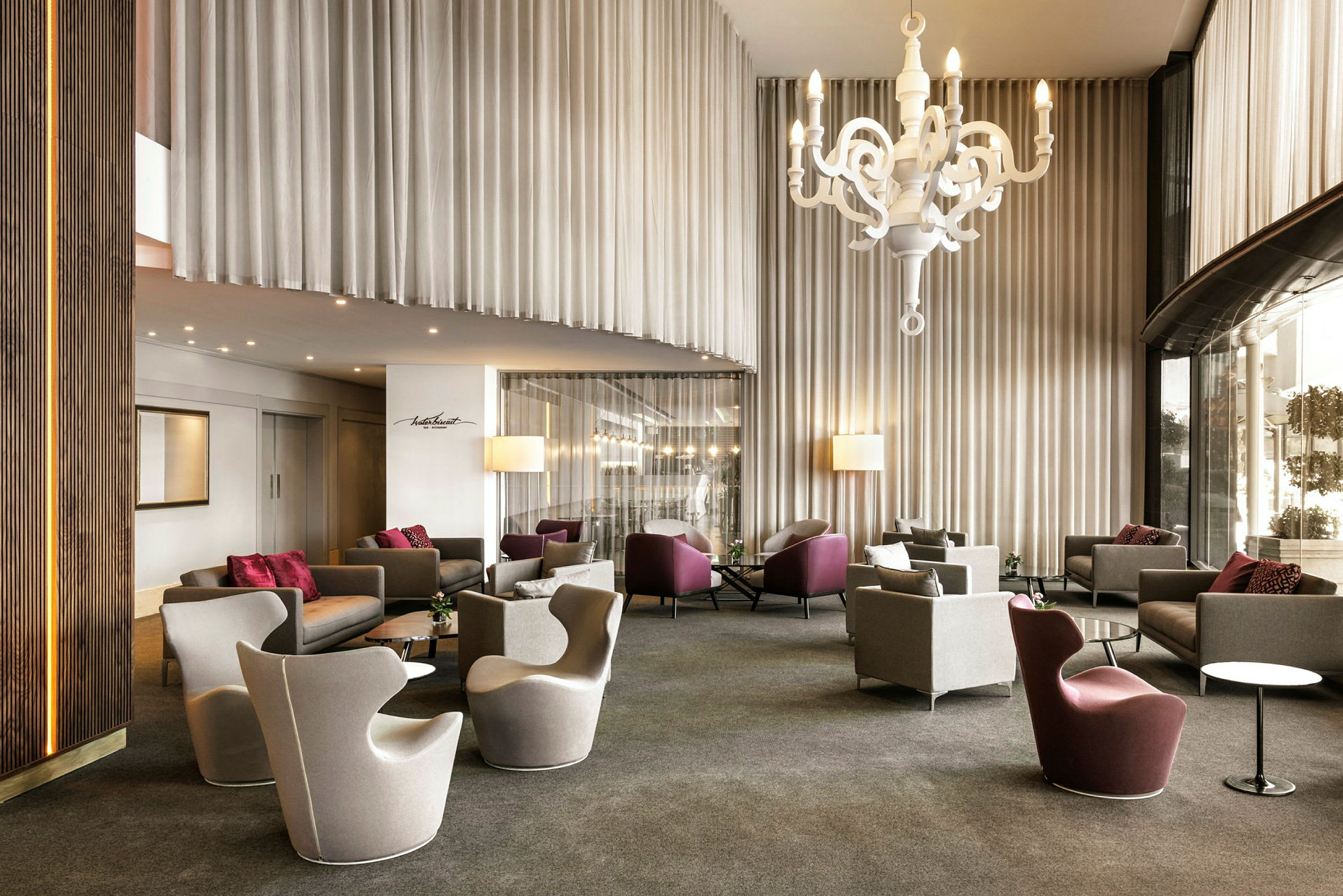 En hotellobby med moderne stoler og sofaer, en stor hvit hengende lysekrone i taket og høye gardiner. (AI-generert tekst)