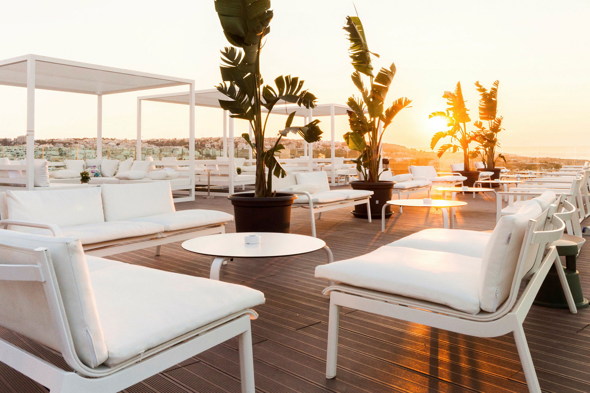 Moderne hvite sofaer og bord på en solfylt terrasse med utsikt over det omkringliggende landskapet, dekorert med store potteplanter. (AI-generert tekst)