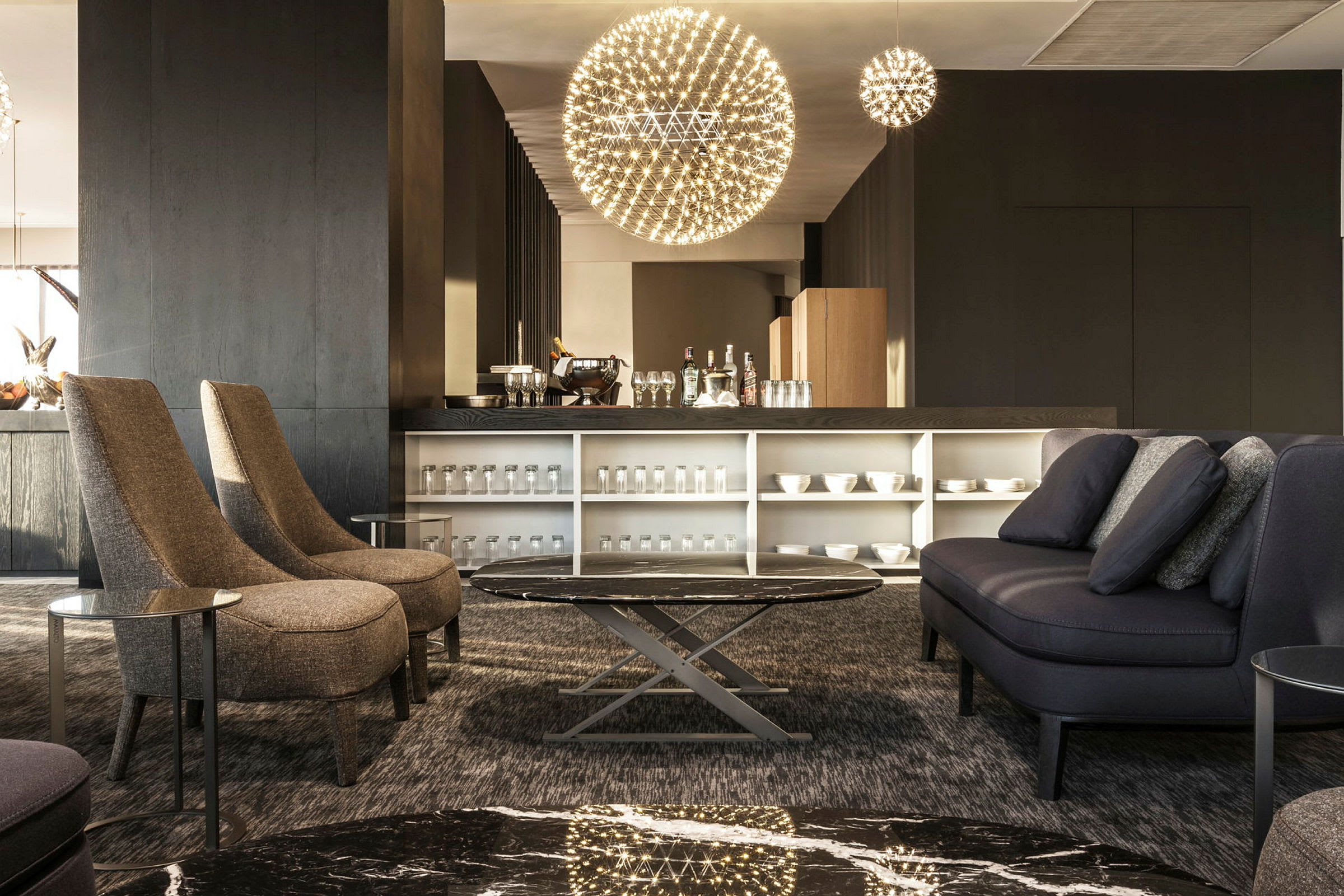 En moderne salong med kaffebar og sittegruppe med sofaer og stoler. Rommet er godt opplyst med unike hengende lamper og har et elegant innredet bord. (AI-generert tekst)