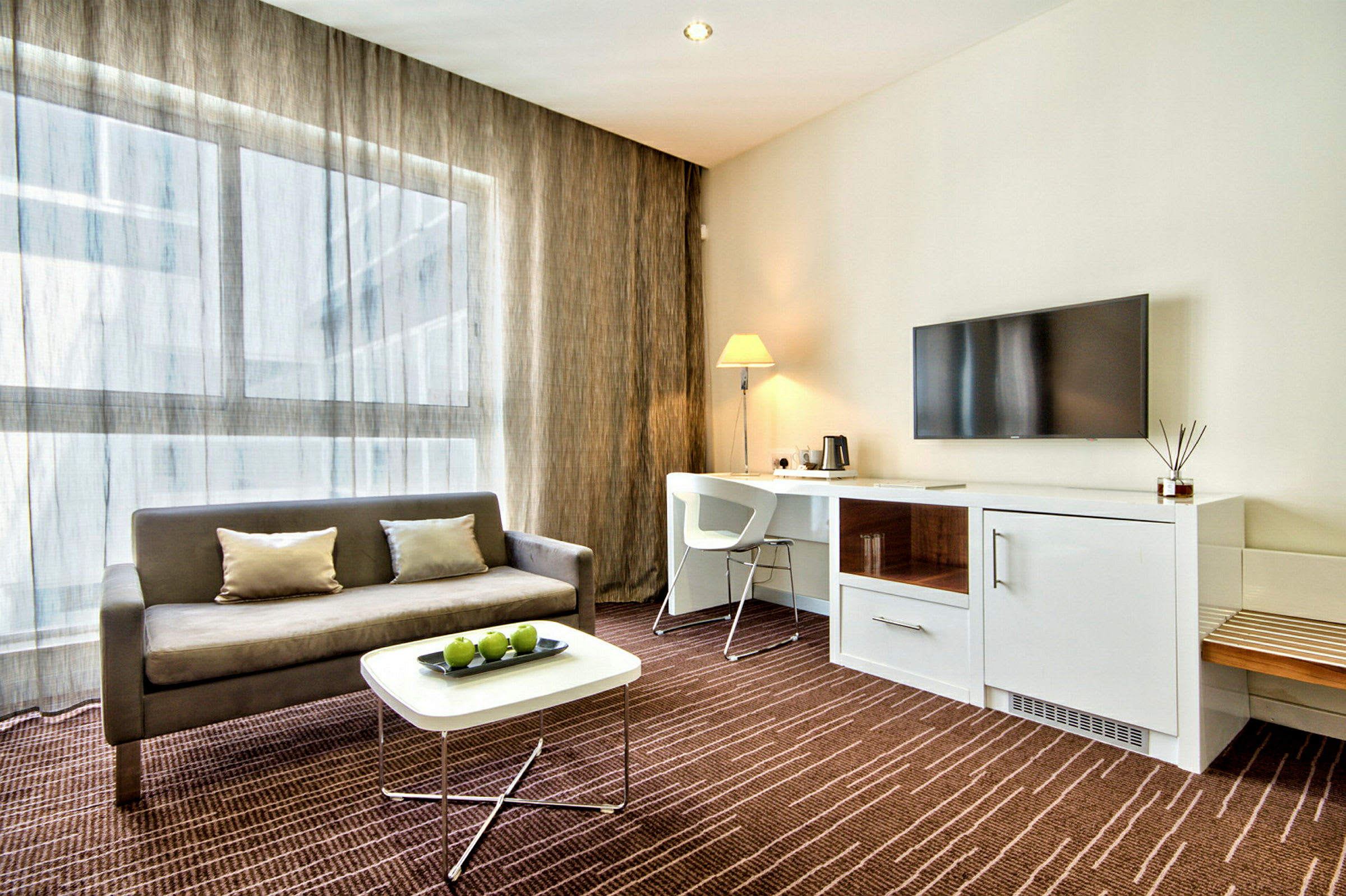Et moderne hotellrom med en sofa, et sofabord med grønne epler, en arbeidsplass med stol og lampe, samt en veggmontert TV. (AI-generert tekst)