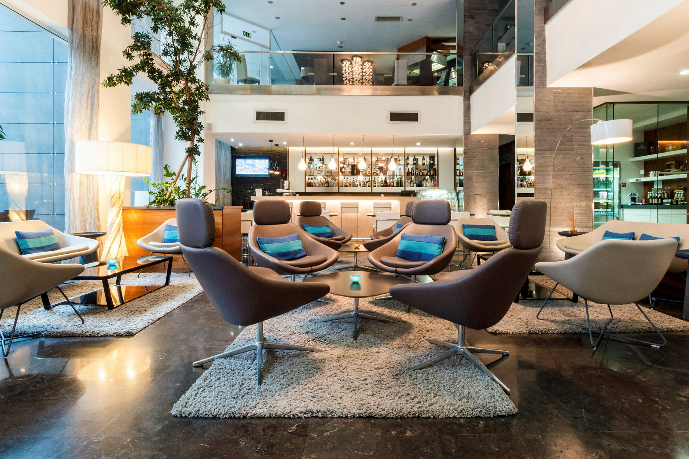 En hotellobby med moderne møbler, flere sofaer og lenestoler med puter i et stilrent miljø. En bar kan sees i bakgrunnen. (AI-generert tekst)
