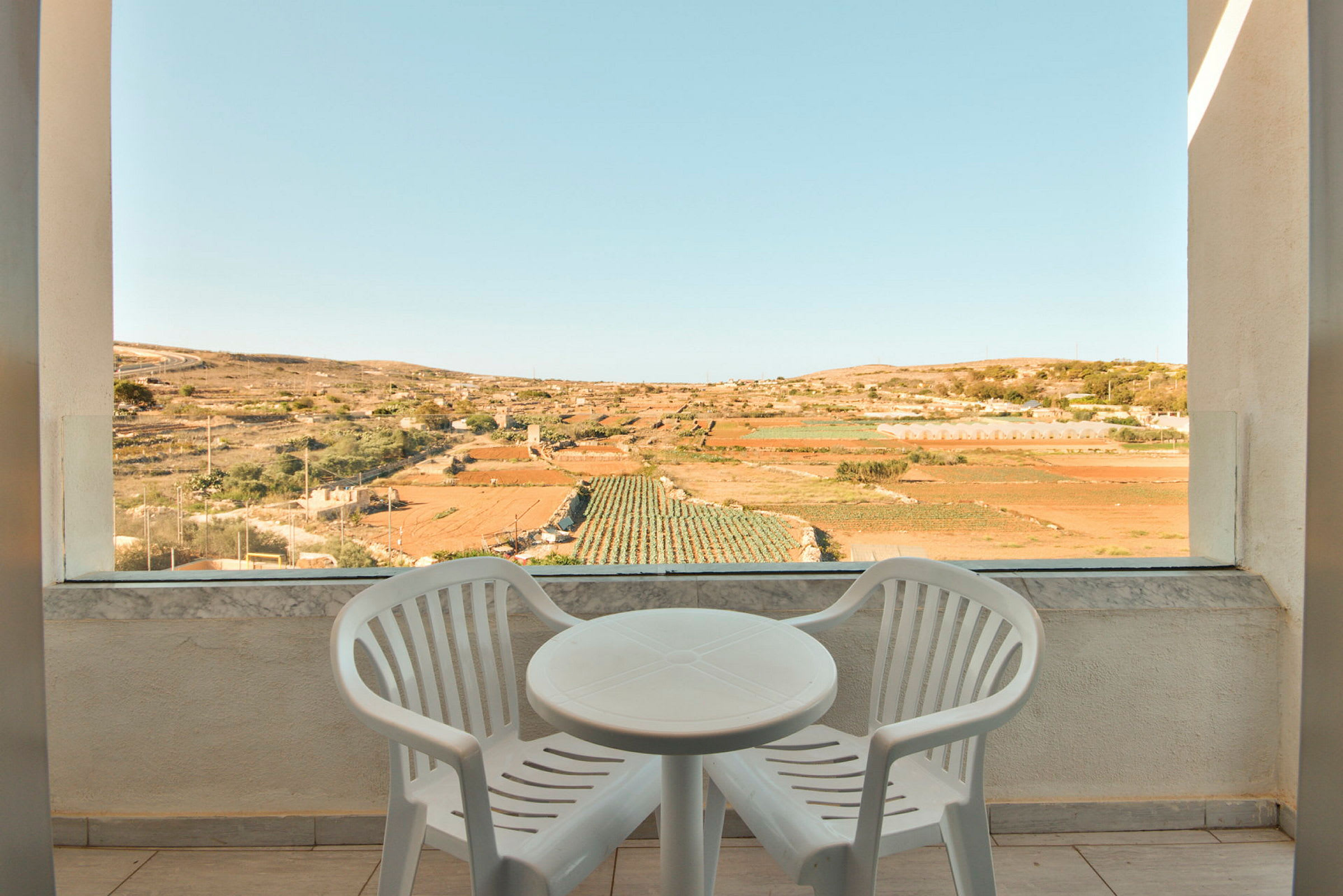 To hvite plaststoler og et rundt bord på en terrasse med utsikt over dyrkingsmarker. (AI-generert tekst)