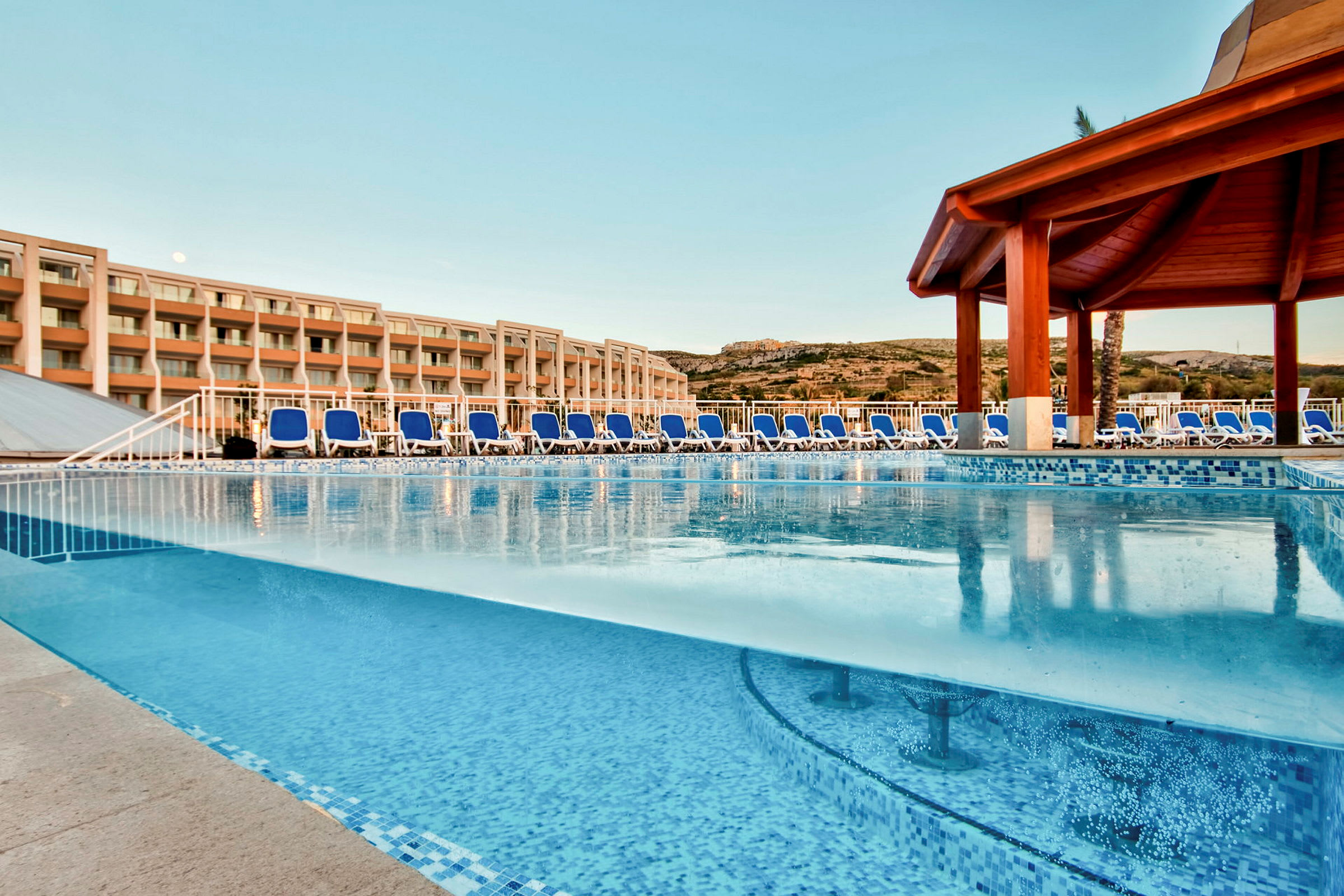 Utendørs svømmebasseng med solsenger og hotellbygning i bakgrunnen. (AI-generert tekst)