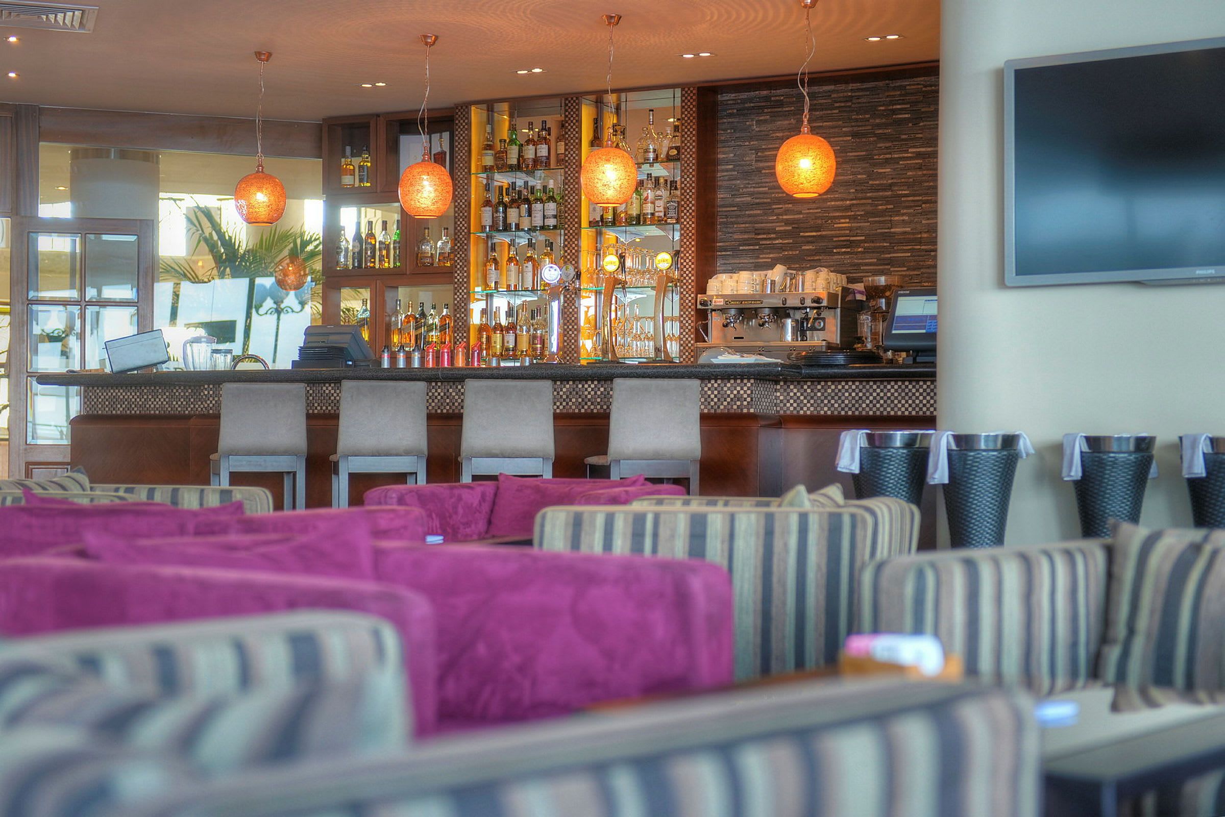 Et interiørbilde av en elegant bar med rader av drikkevarer, pene barkrakker og moderne lamper. I forgrunnen sees sitteplasser med fargede puter. (AI-generert tekst)