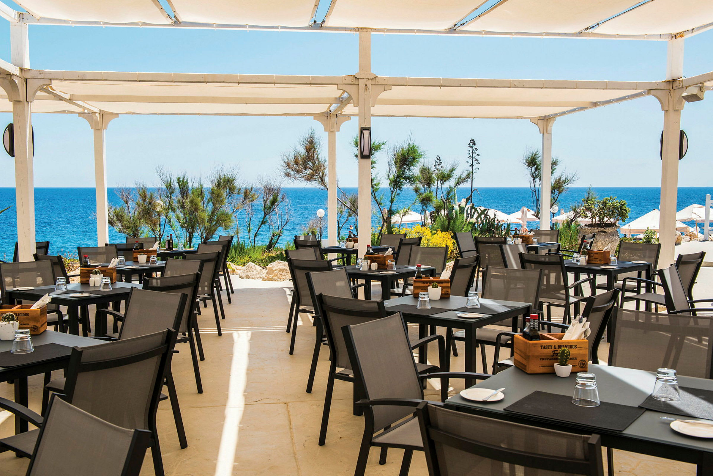 En terrasse med uteservering, bord og stoler under et hvitt tak, med havet og vegetasjon i bakgrunnen. (AI-generert tekst)