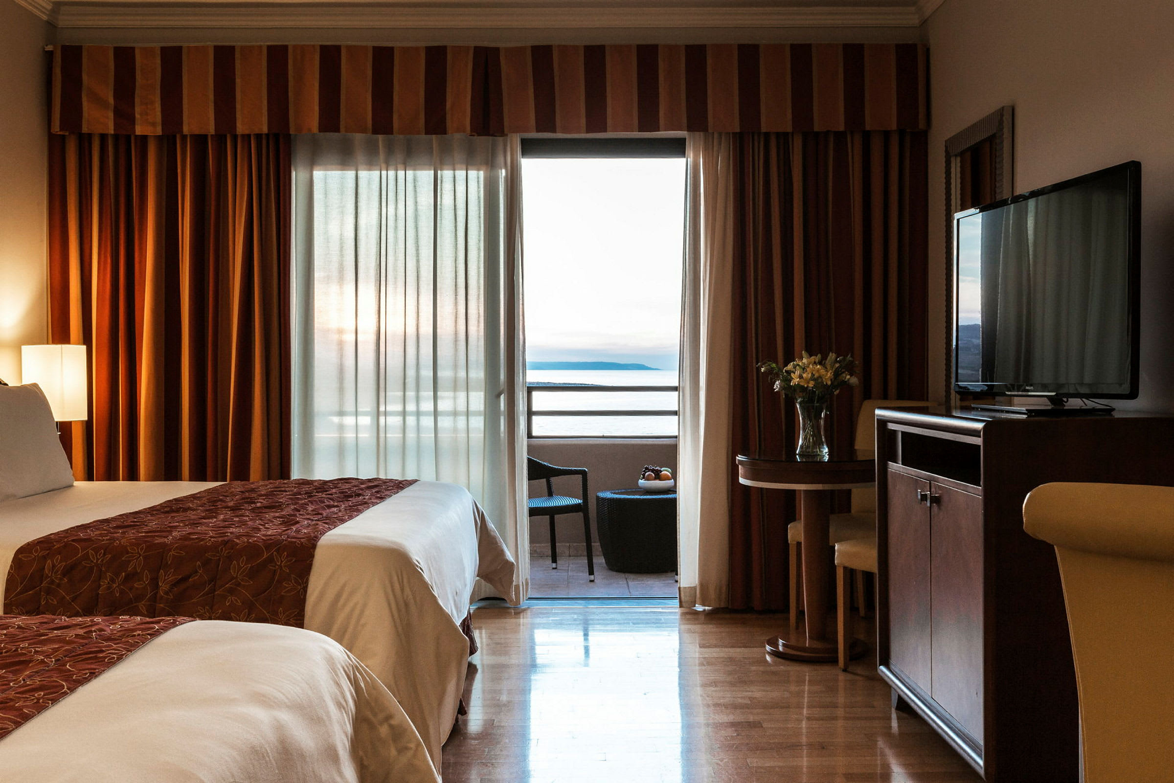 Et hotellrom med to senger, en balkong med utsikt over havet, og et mindre bord med stoler. (AI-generert tekst)