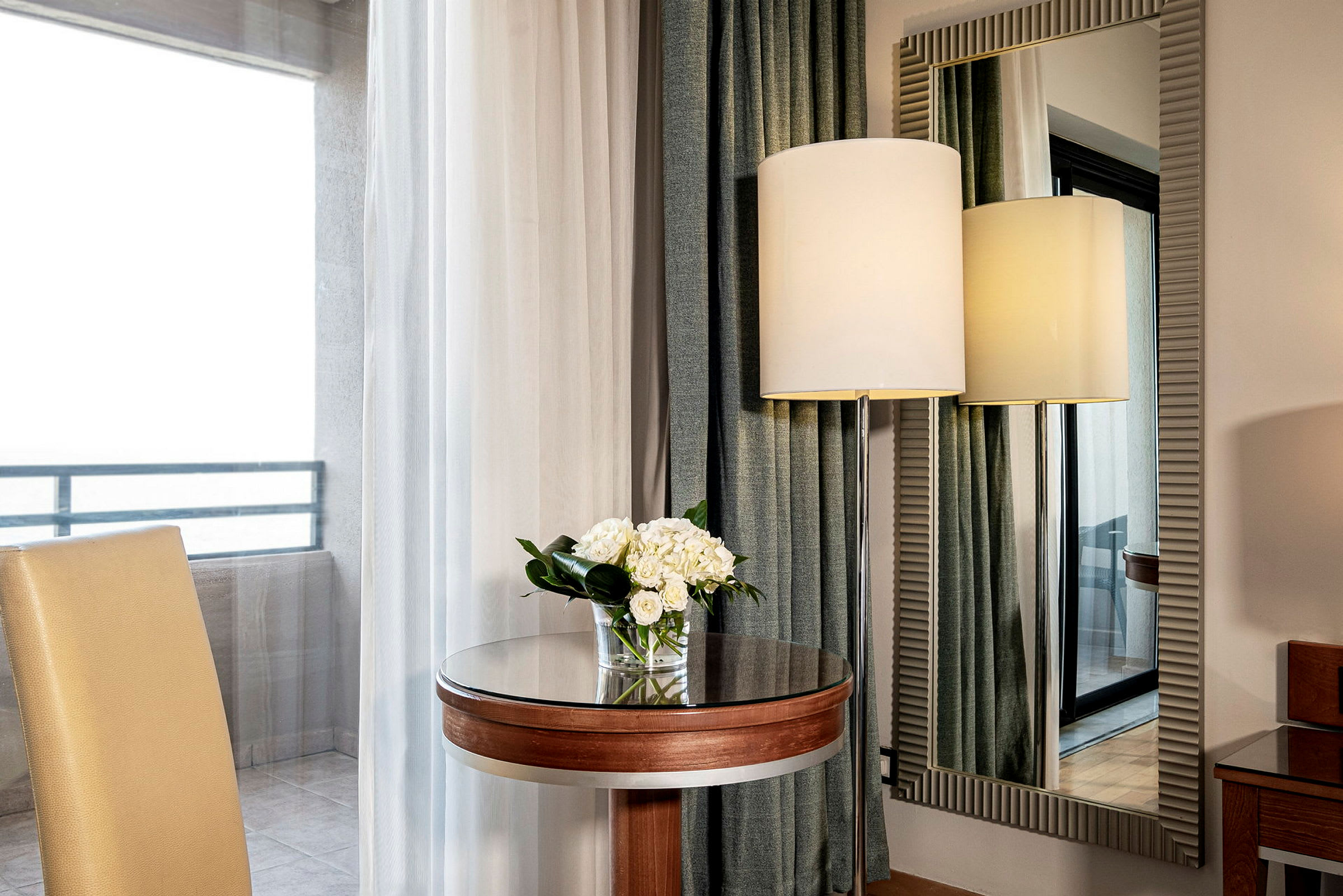 Et elegant hotellområde med en stol, et lite rundt bord med en blomsterbukett, et stort speil og en gulvlampe sett gjennom et vindu. (AI-generert tekst)