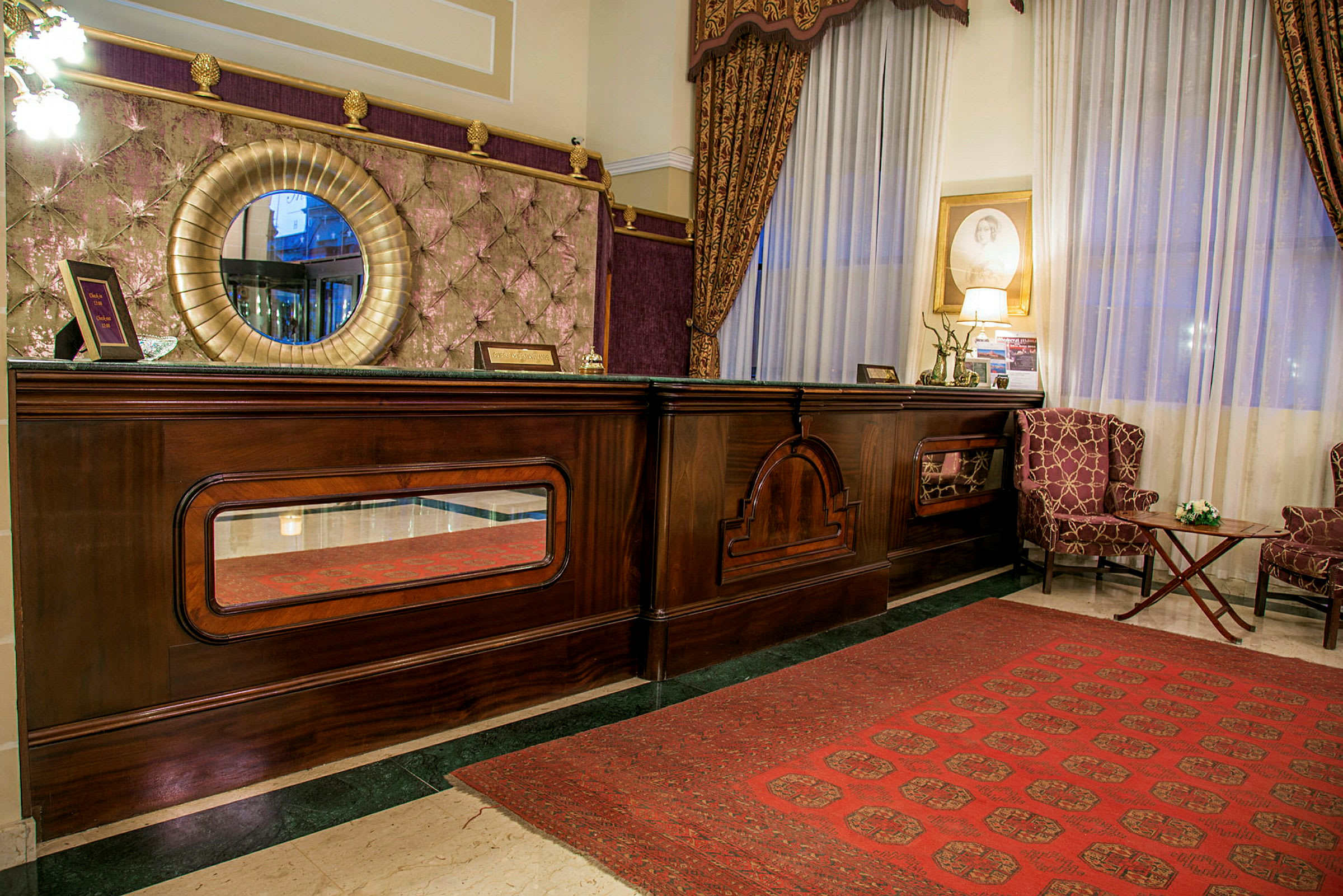 Bilde av en hotellresepsjon med en elegant trekke, rundt speil og to lenestoler foran et stort vindu. (AI-generert tekst)