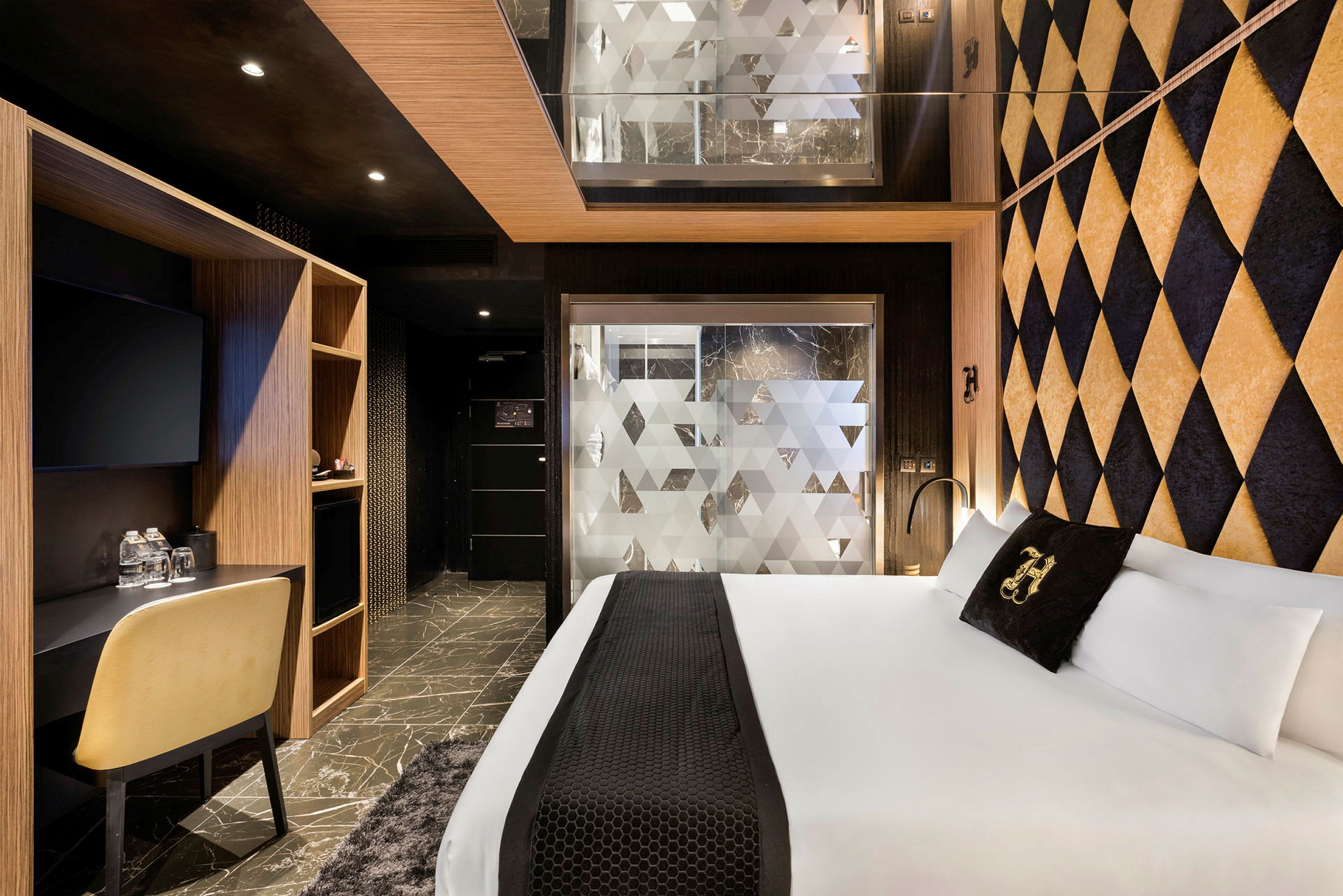 Et hotellrom med moderne interiør. Rommet har en stor seng, geometriske dekorvegger og et skrivebord med stol. En frostet glassvegg skiller badet fra sovedelen. (AI-generert tekst)