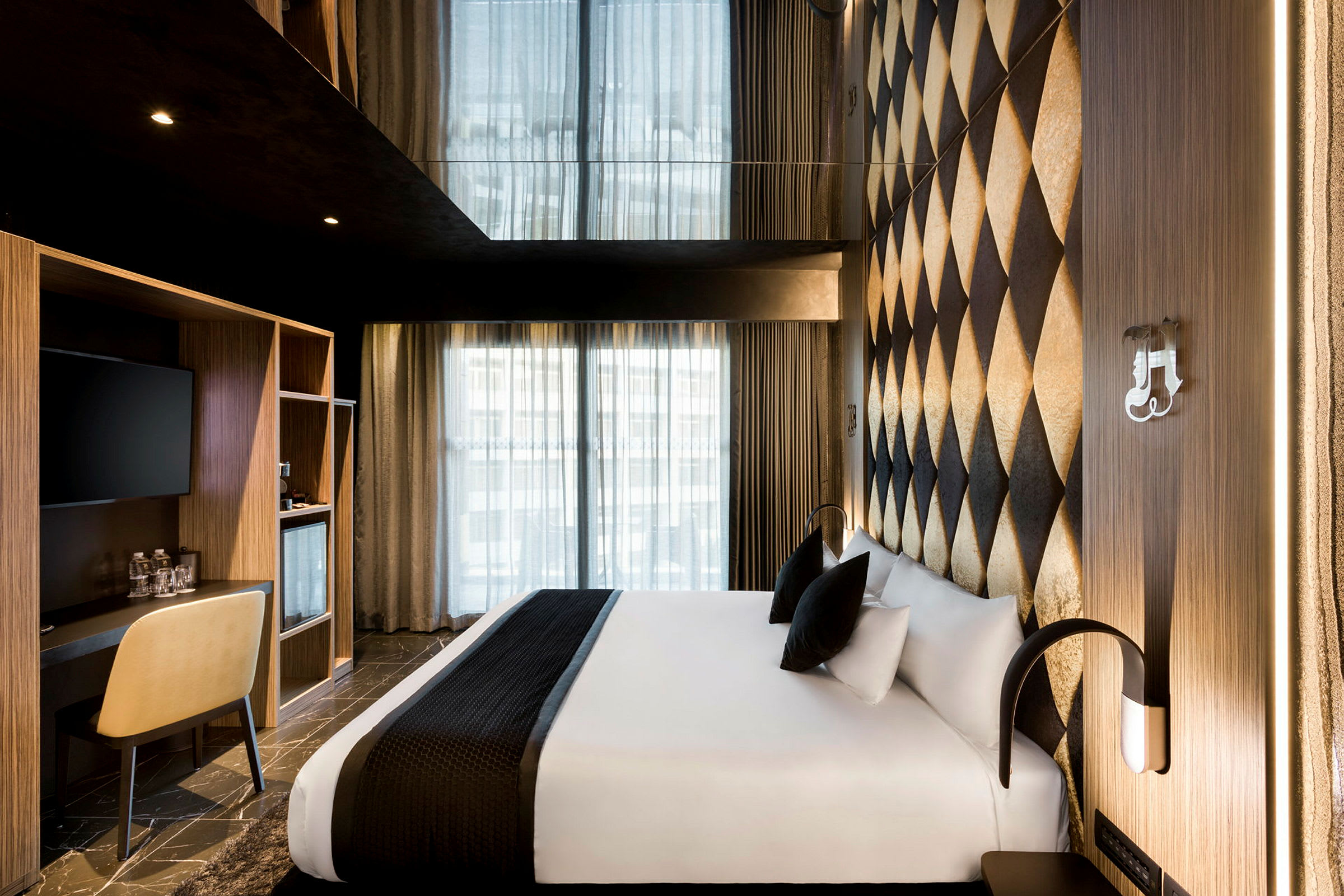 Et moderne hotellrom med en bred seng dekket med hvitt sengetøy og sorte puter. Veggene har et dekorativt rutemønster, og ved siden av sengen er det et lite skrivebord med en stol. (AI-generert tekst)