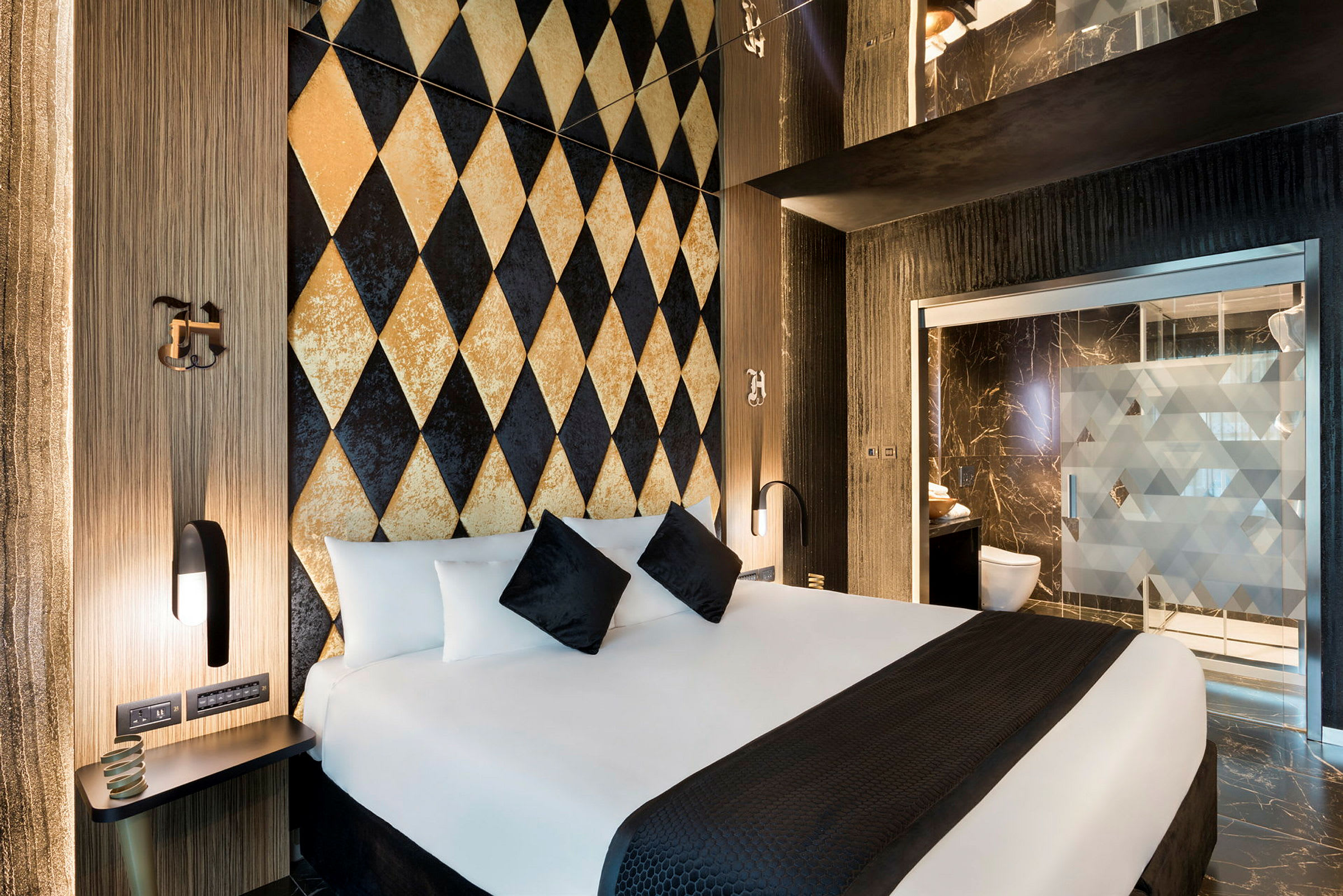 Et moderne hotellrom med dekorvegg i harlekinmønster og en stor seng med hvite og svarte puter. Badet ses i bakgrunnen. (AI-generert tekst)