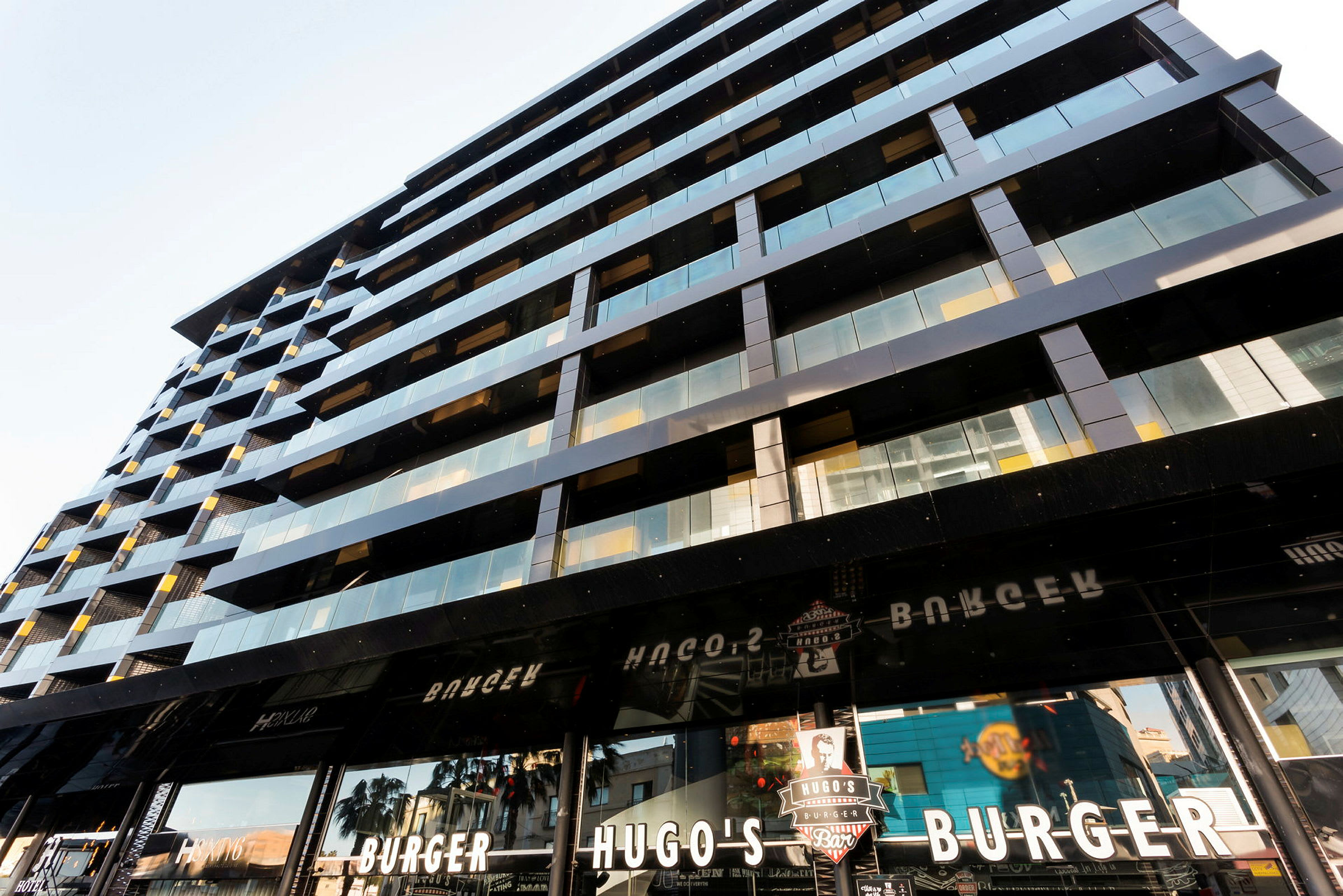 En moderne bygning med glass- og metallfasade samt skilt for en burgerrestaurant kalt Hugo's Burger. (AI-generert tekst)