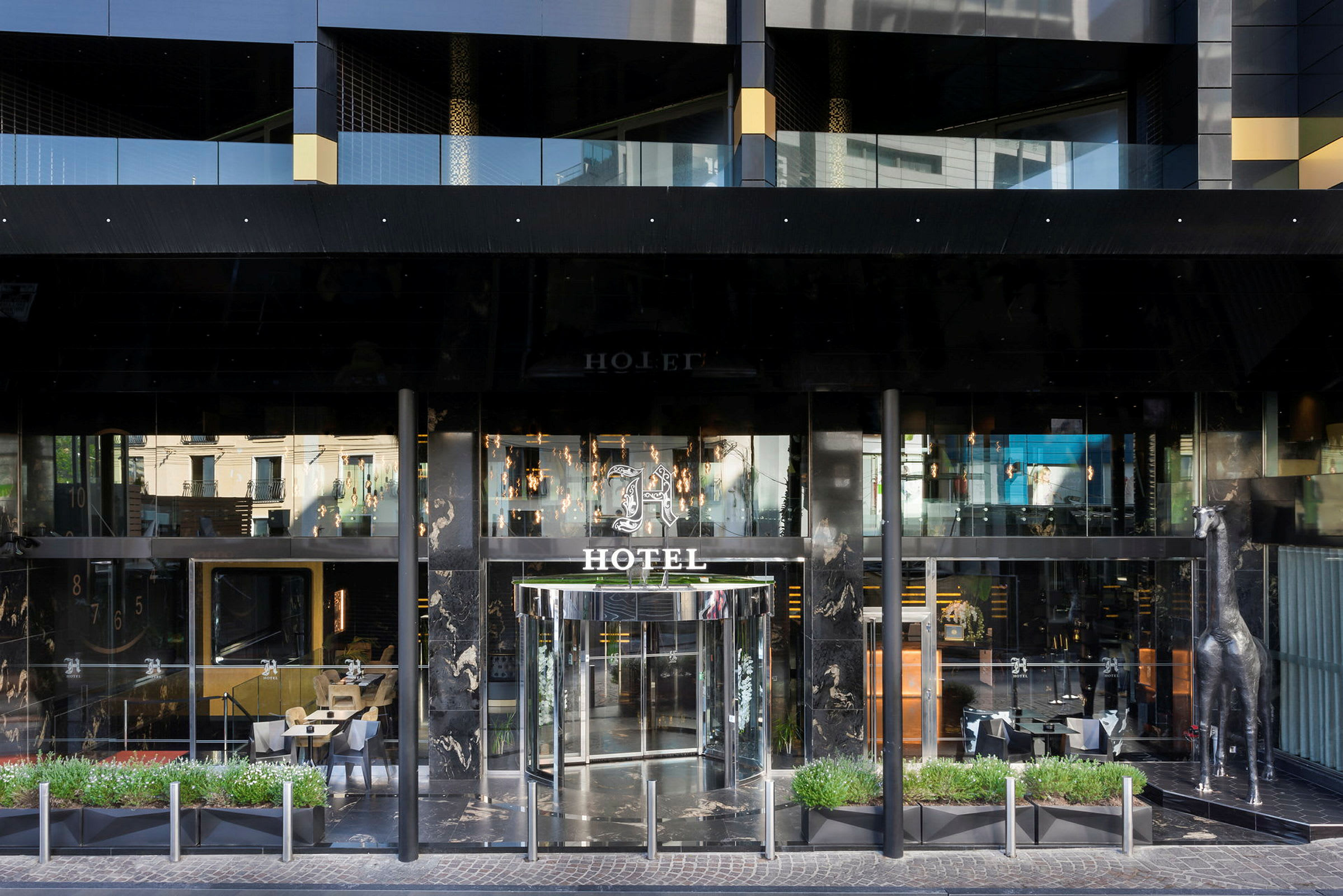 Forsiden av et hotell med glassdører og en dekorativ sølvfarget sjiraffstatue ved siden av inngangen. (AI-generert tekst)