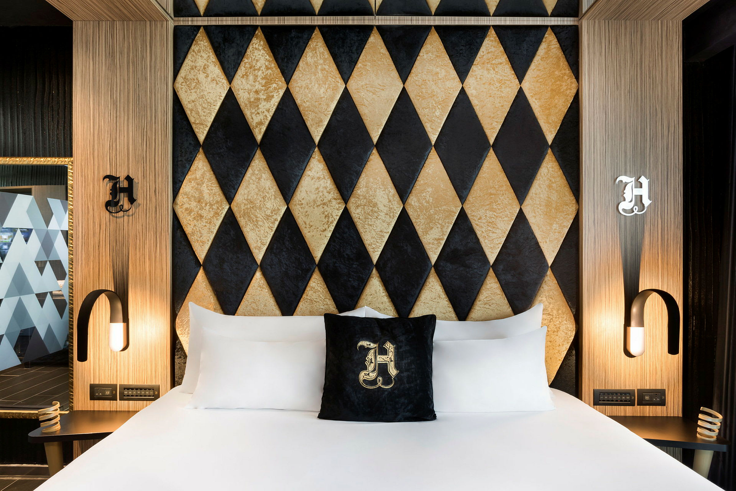 Et hotellrom med en seng med hvite laken og en dekorativ pute med bokstav. Bak sengen er en vegg med diamantmønster i svart og gull. (AI-generert tekst)