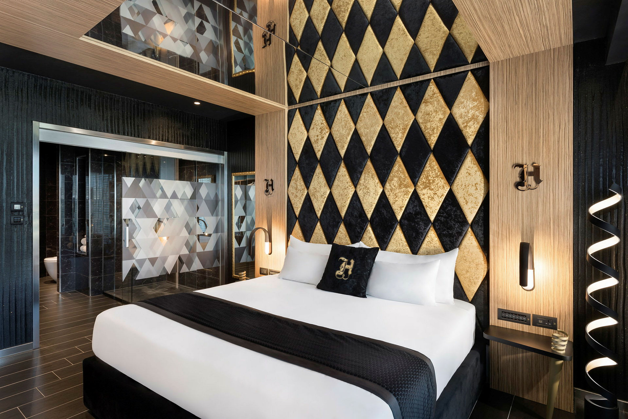 Et hotellrom med elegant design. En dobbeltseng med hvite laken og et svart pledd er sentralt plassert. Bakgrunnen er en vegg med gullfargede og svarte diamantmønstre. Det er separate glassdører med geometriske detaljer som fører til et bad. (AI-generert tekst)