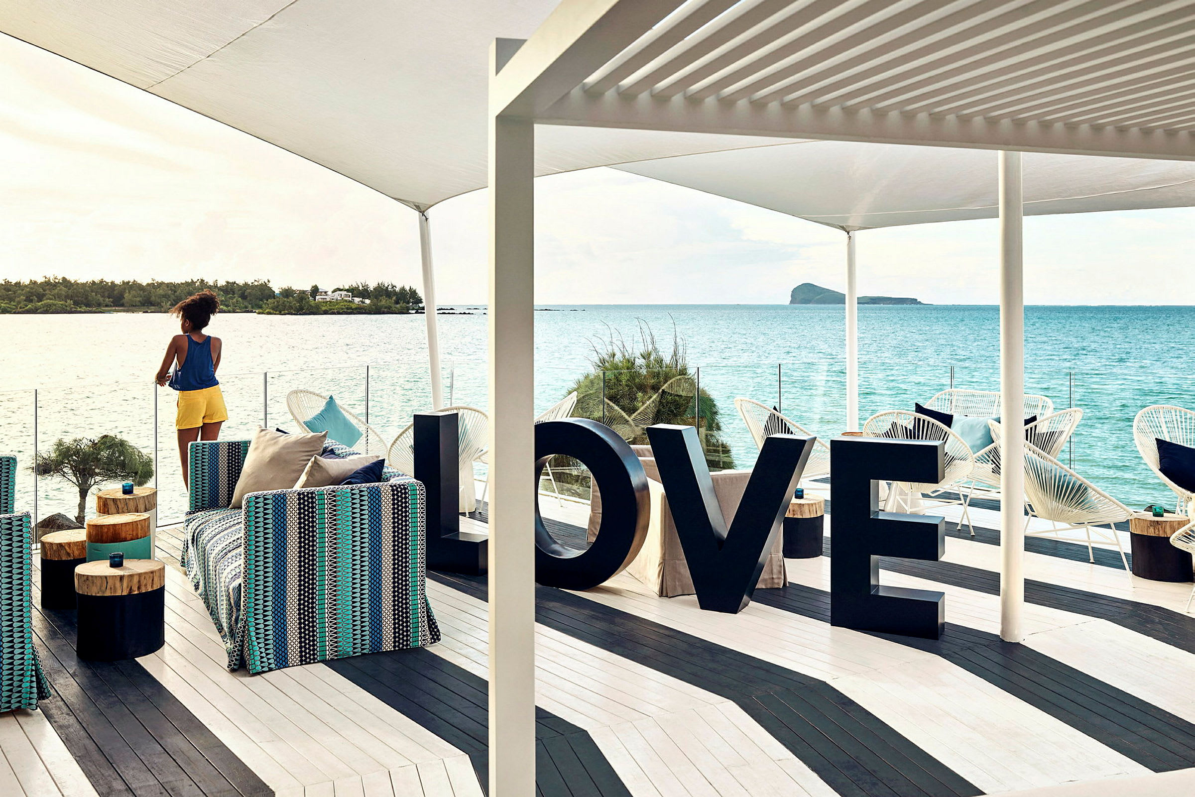 En kvinne står på en terrasse med utsikt over havet, der det finnes møbler og store bokstaver som danner ordet 'LOVE'. (AI-generert tekst)