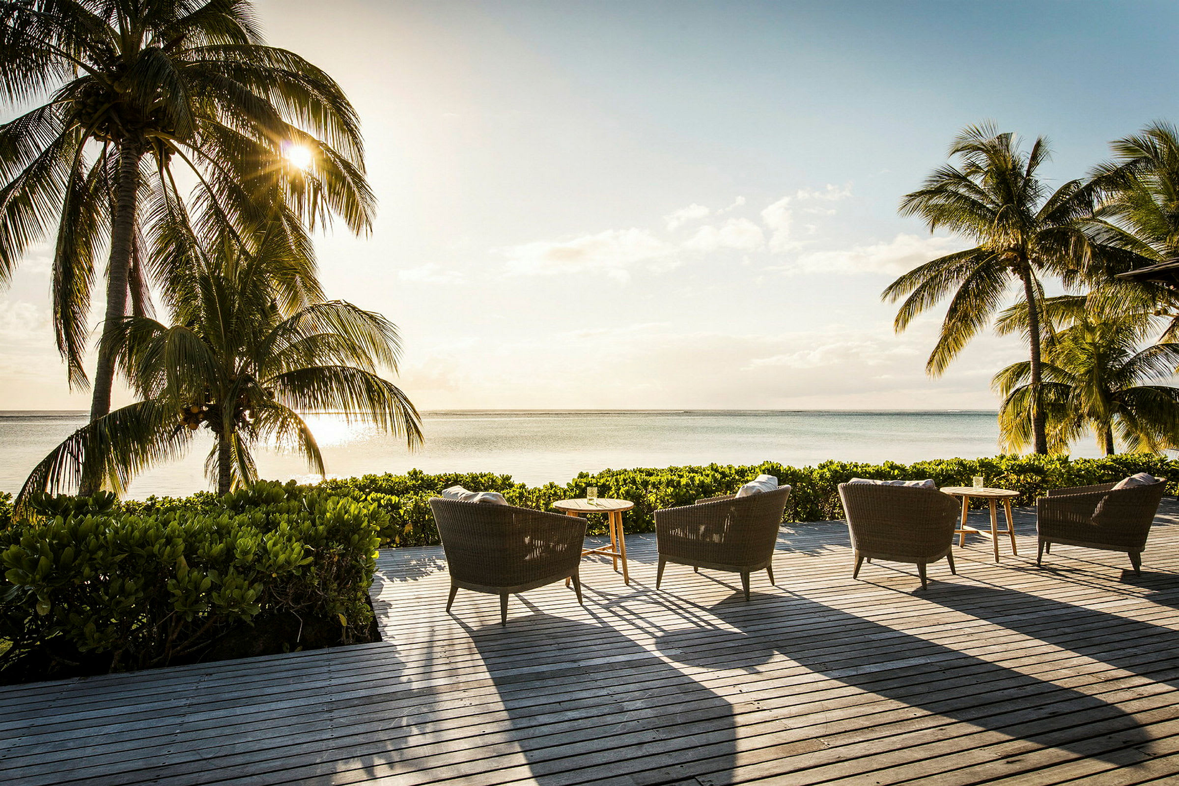 Utemøbler på en terrasse med utsikt over havet og omkringliggende palmer i solnedgangen. (AI-generert tekst)
