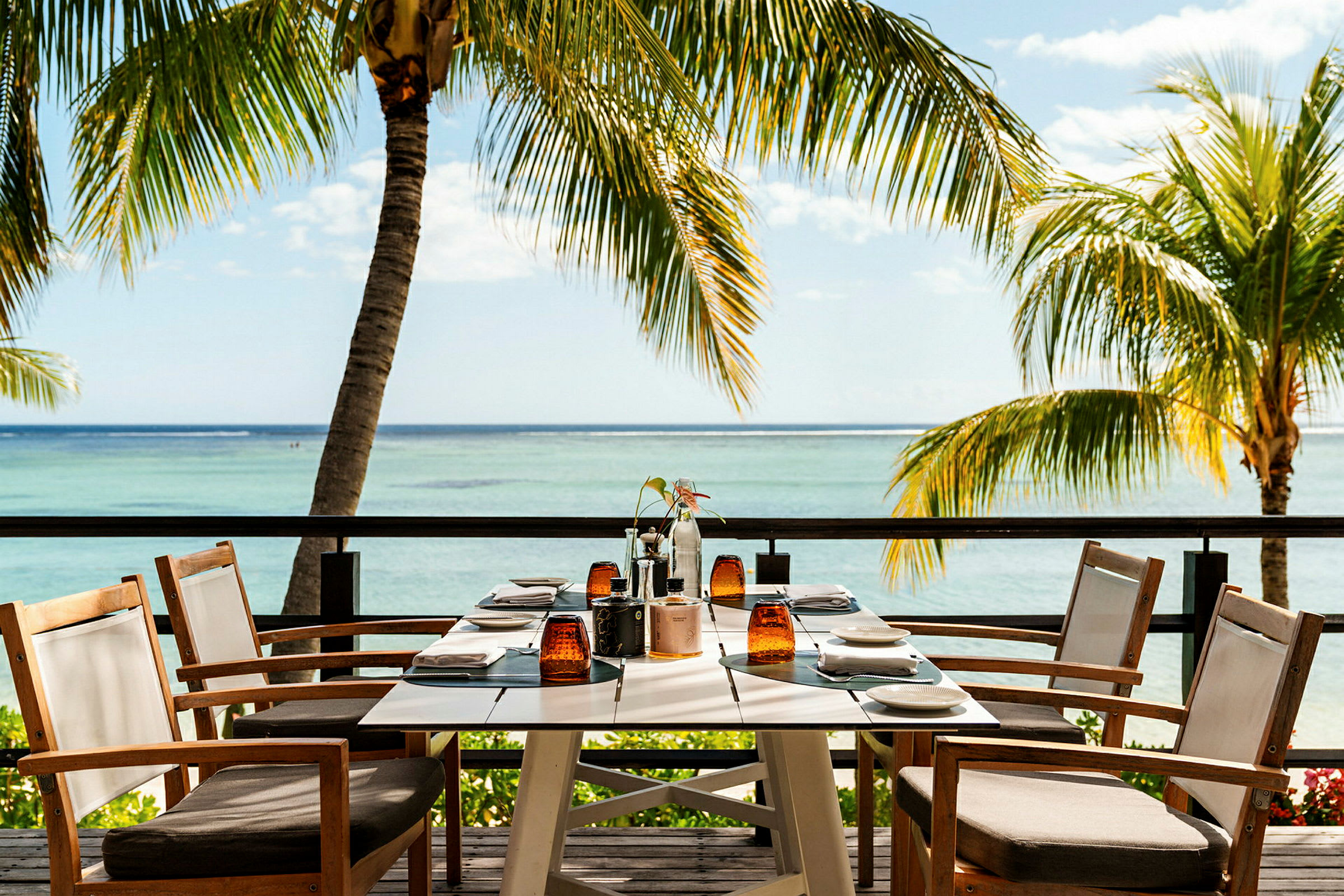 Et dekket bord med sitteplasser står på en terrasse med utsikt over havet og omkringliggende palmer. (AI-generert tekst)