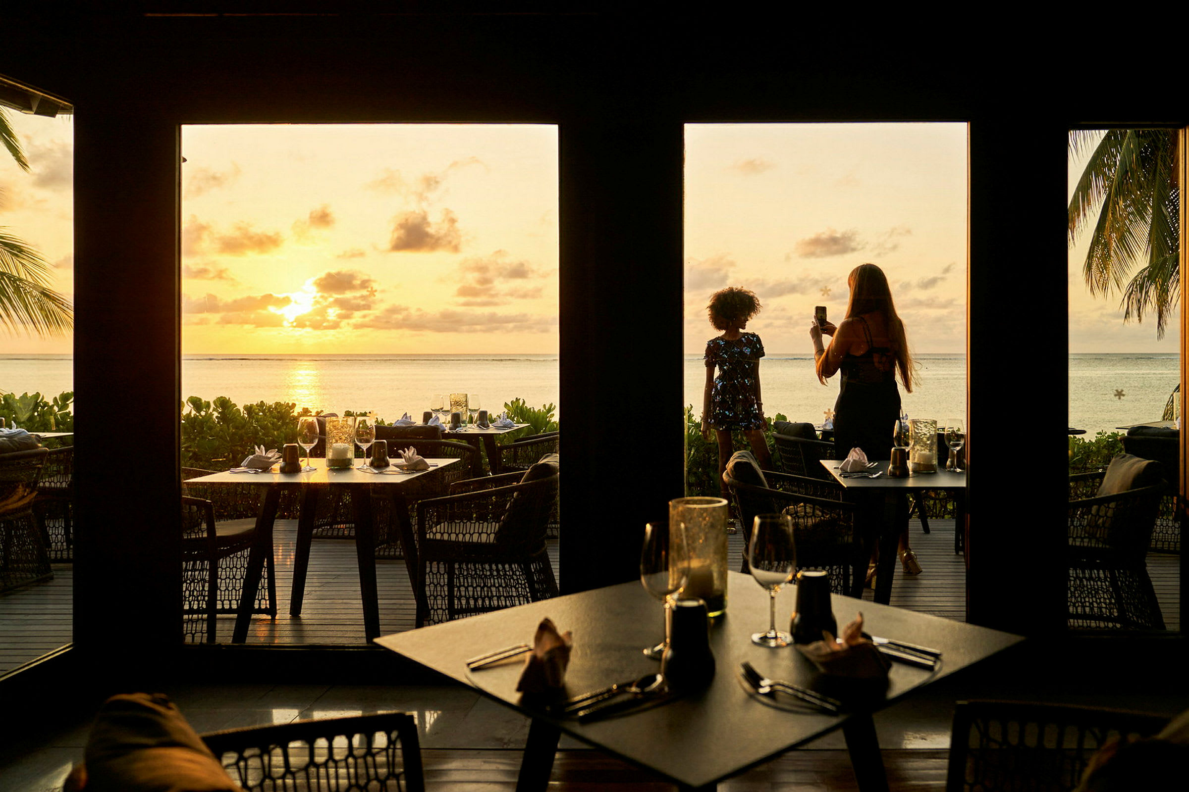 Bildet viser en restaurant med utsikt over en strand ved solnedgang, med bord plassert både inne og ute, samt palmer og havet i bakgrunnen. (AI-generert tekst)