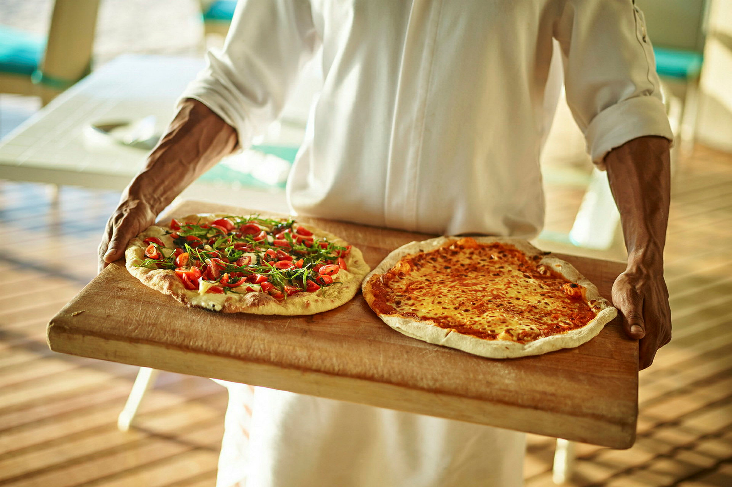 En person bærer to pizzaer, en med friske tomater og ruccola og en med smeltet ost, på et trefat. (AI-generert tekst)