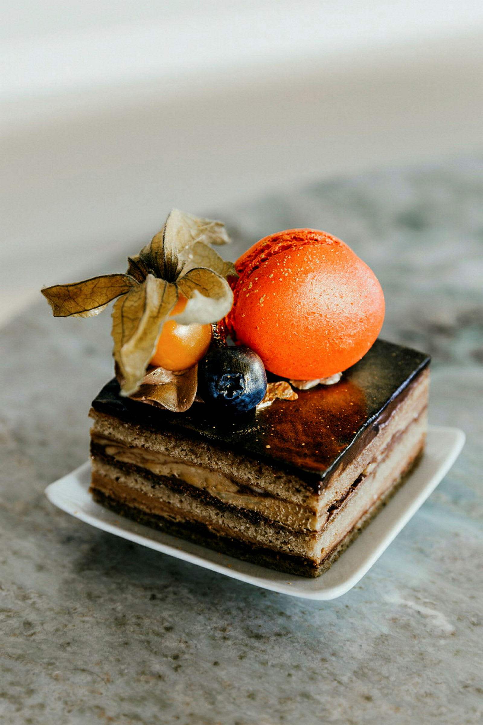 Nærbilde av en elegant dessert med sjokoladeglasur, lag med krem, dekorert med en oransje makron, en physalis og ett blåbær. (AI-generert tekst)