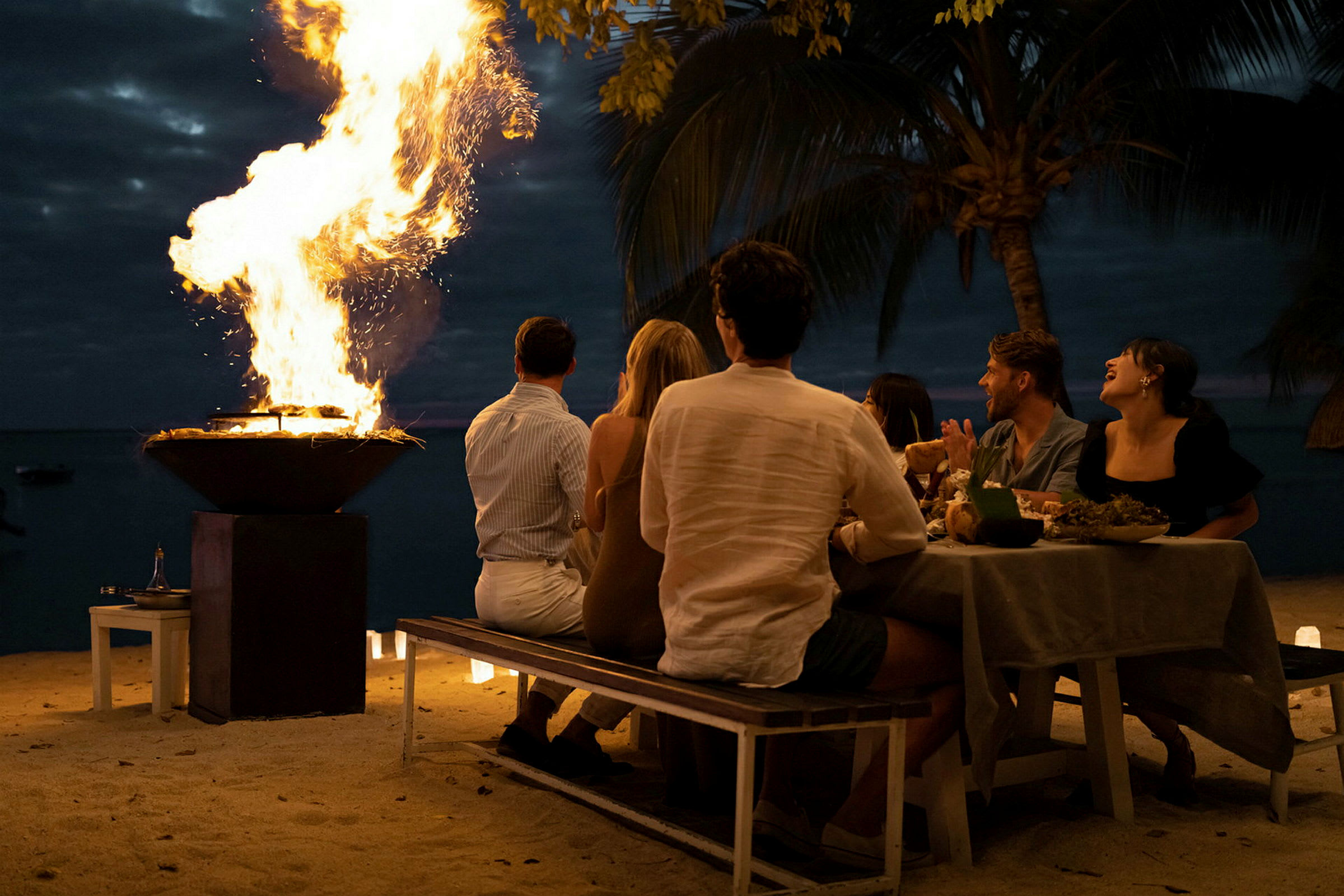En gruppe mennesker sitter ved et dekket bord på stranden nær en ildbolle med flammer om kvelden. (AI-generert tekst)