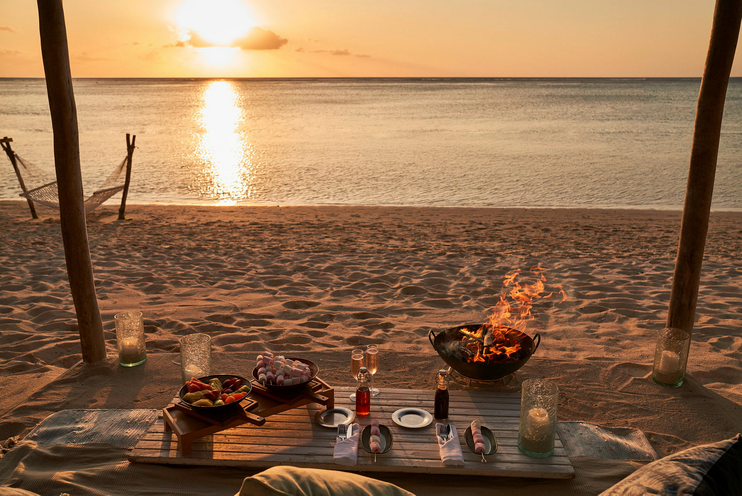 Dekket bord på en strand med en solnedgang i bakgrunnen og et bål i en ildsted. (AI-generert tekst)