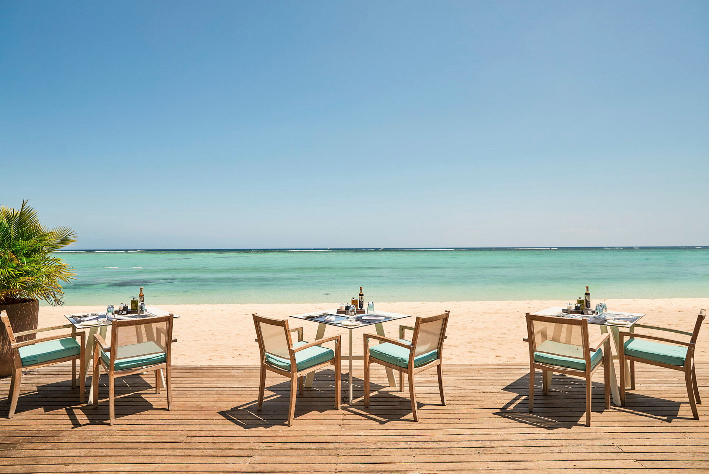 Bildet viser en treterrasse med bord og stoler, plassert nær en sandstrand med utsikt over havet. (AI-generert tekst)