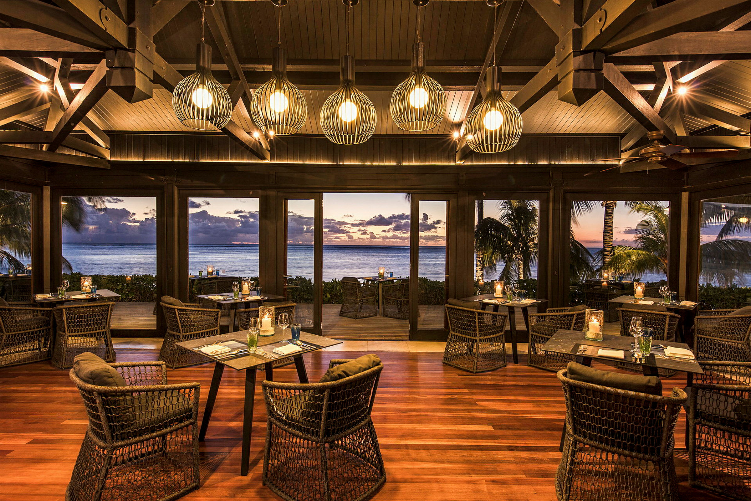 En elegant restaurant med treinteriør og moderne stoler. Vinduene gir utsikt over havet med en solnedgang i det fjerne. (AI-generert tekst)