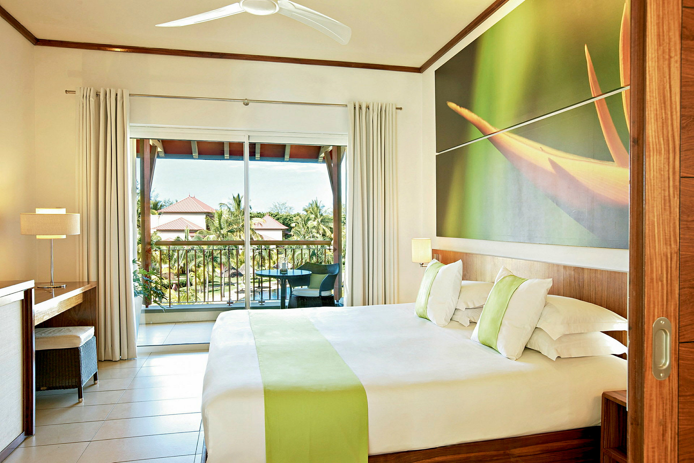 Hotellrom med en dobbeltseng, en stor vindusvegg og en balkong med utsikt over grønne områder og takene på nærliggende bygninger. (AI-generert tekst)