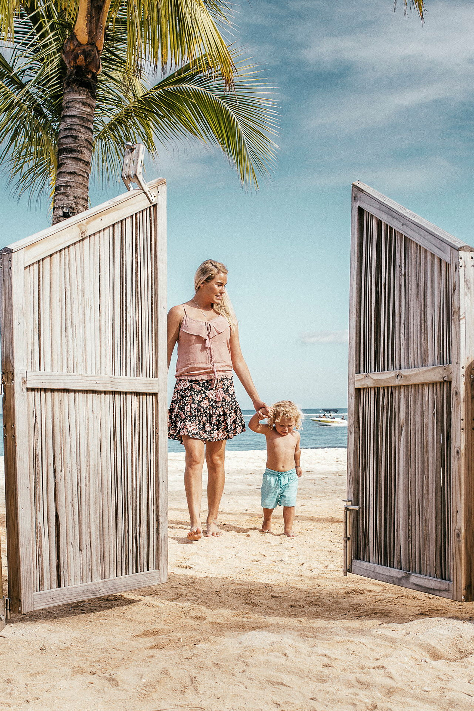 En person i sommerklær og et barn går hånd i hånd gjennom en tredør mot en sandstrand. (AI-generert tekst)