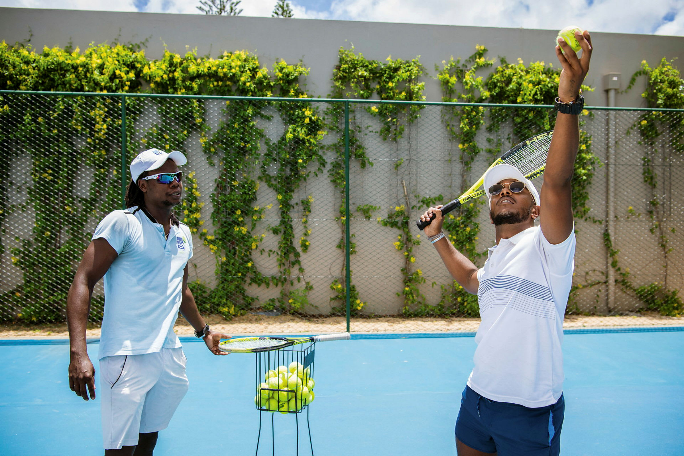 To personer spiller tennis på en utendørs tennisbane med en beholder fylt med tennisballer. (AI-generert tekst)