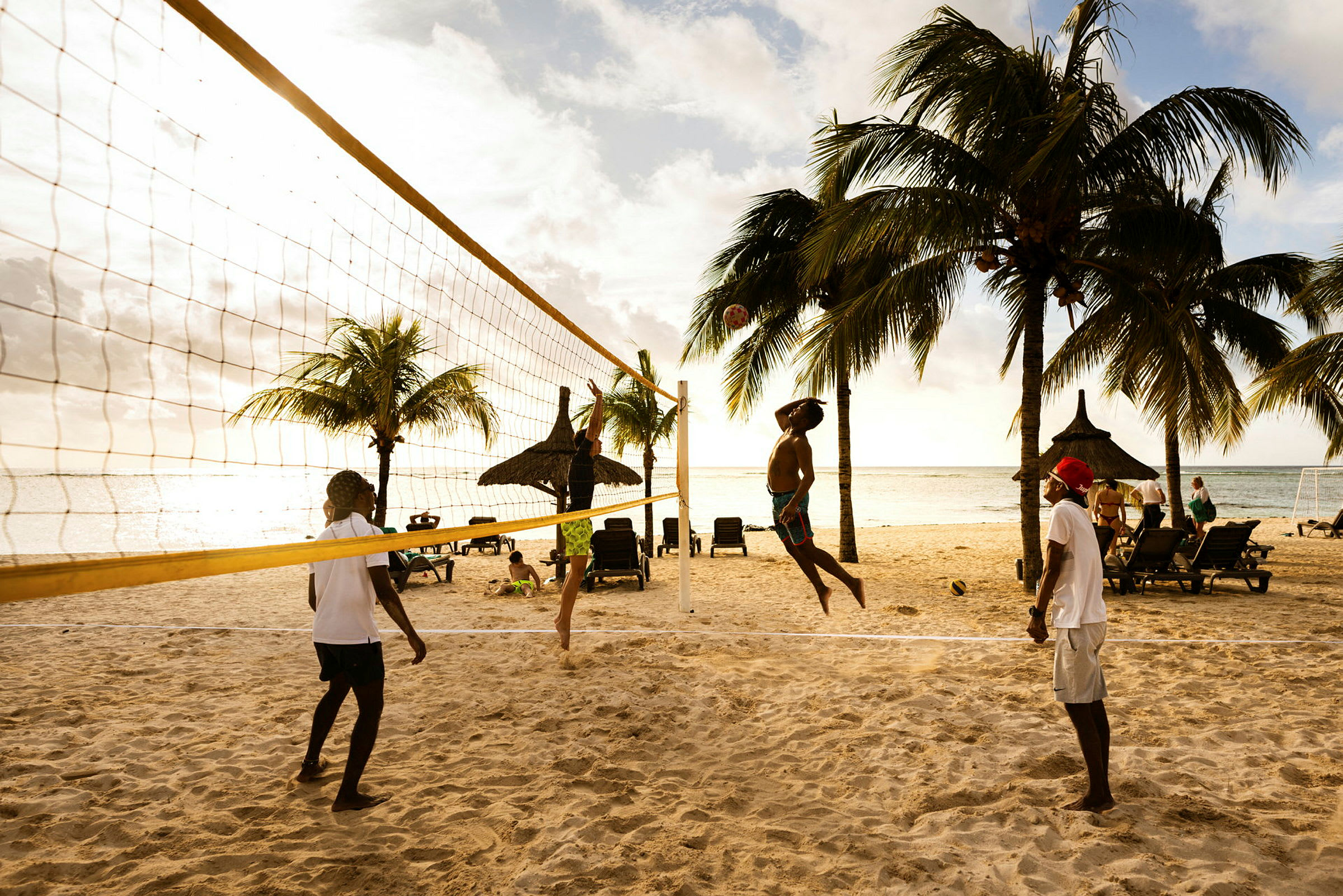 Fire personer spiller sandvolleyball på en strand med solsenger og palmer i bakgrunnen. (AI-generert tekst)