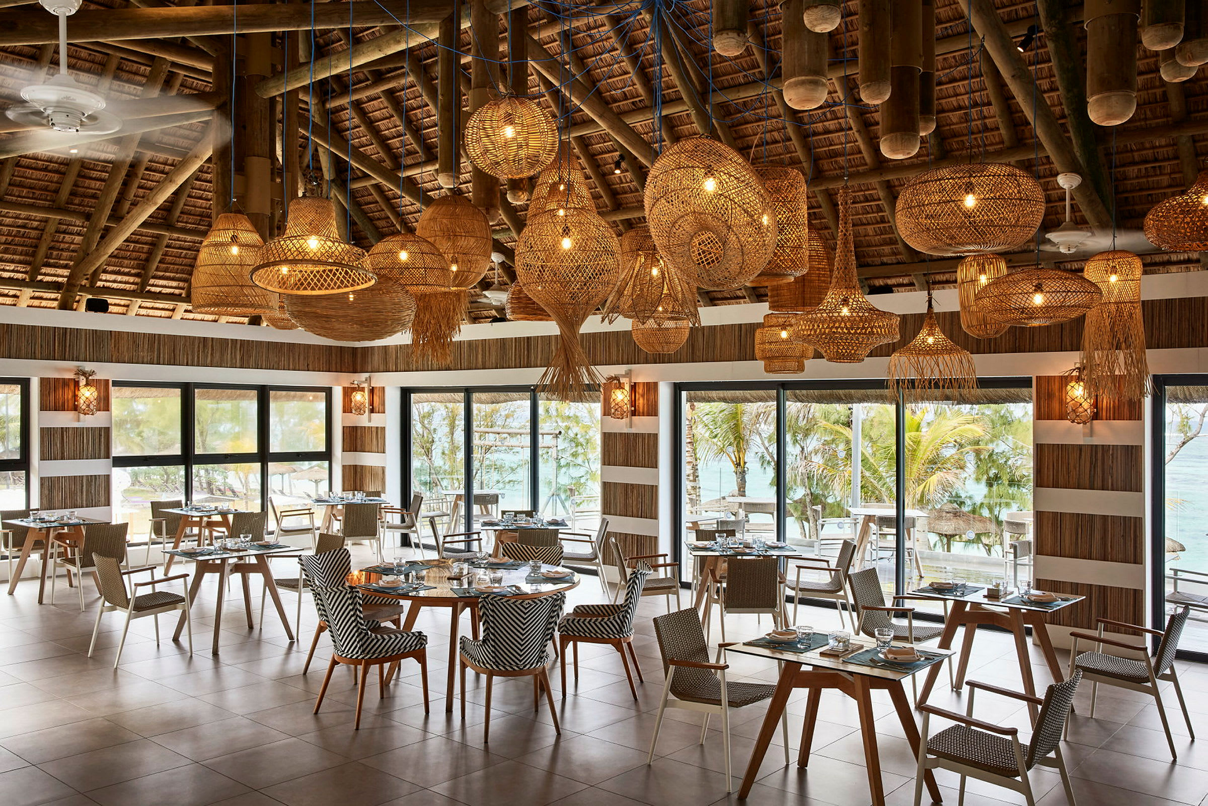 En restaurant med bord og stoler, store vinduer med utsikt mot tropisk miljø og dekorative taklamper. (AI-generert tekst)